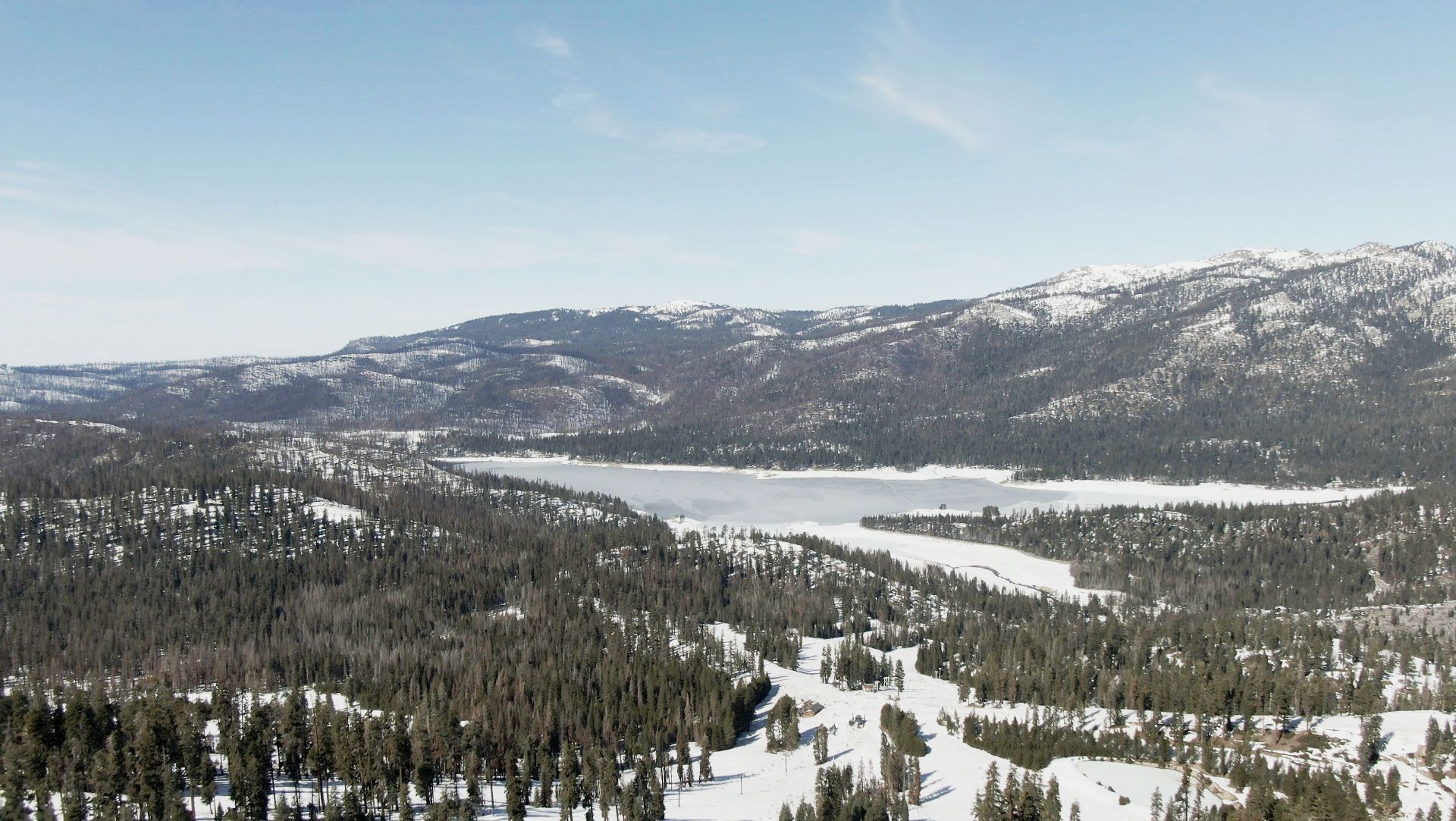 Snowy mountain landscape with frozen lake and evergreen trees under a blue sky.