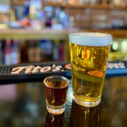 A pint of beer and a shot of liquor on a bar with a Tito's bar mat in the background.