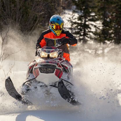 Person riding a snowmobile through snow. They wear an orange jacket, blue helmet, and goggles, in a snowy forest.
