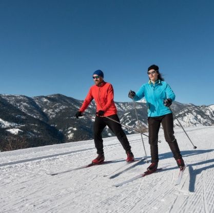 Two people cross-country skiing on a snowy slope under a blue sky, mountains in the background.