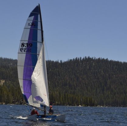 Catamaran sailing on a lake with purple and white sail, trees in the background. Two people are aboard.