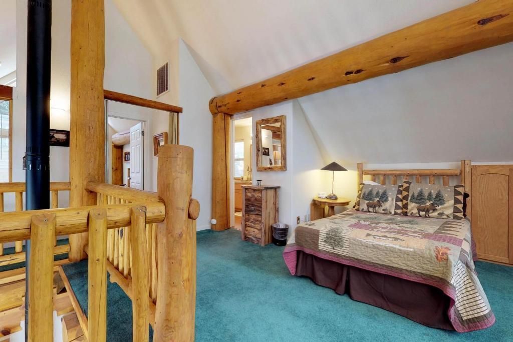 Cozy attic bedroom with log beams, teal carpet, floral bedspread, lamp, and wooden dresser