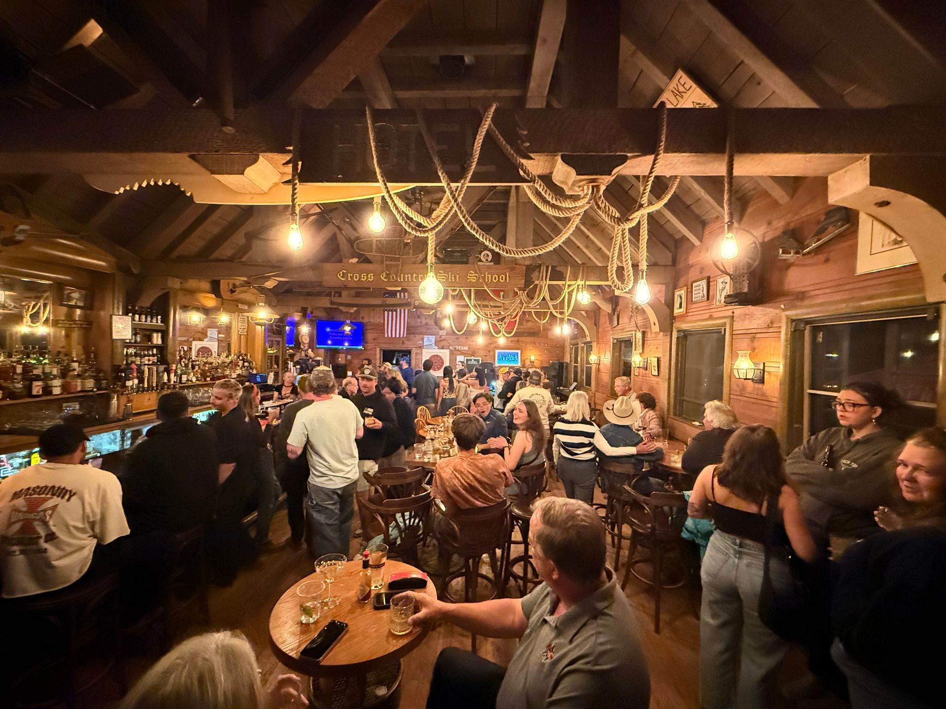 Crowded rustic bar with patrons mingling under warm lights and exposed wooden beams