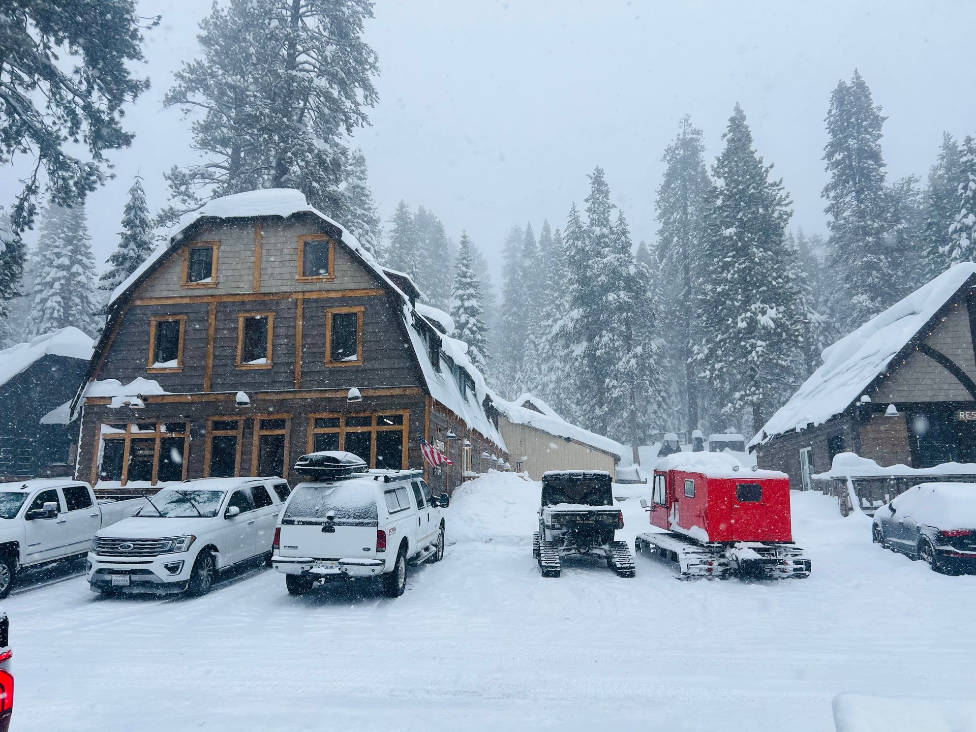 Snowy mountain lodge parking lot with several cars and a red snowmobile in front of cabins