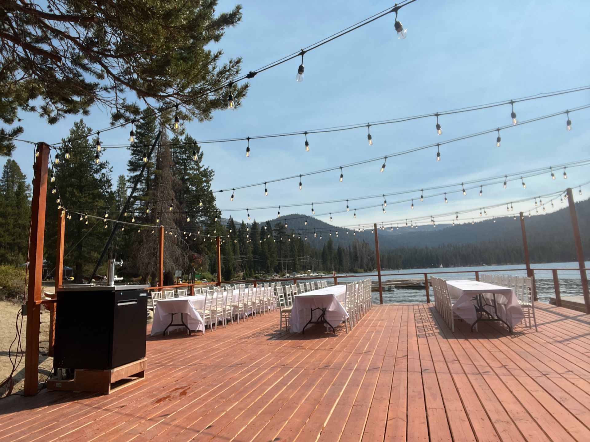 Outdoor deck with white tables, string lights, and mountain lake view under a clear blue sky