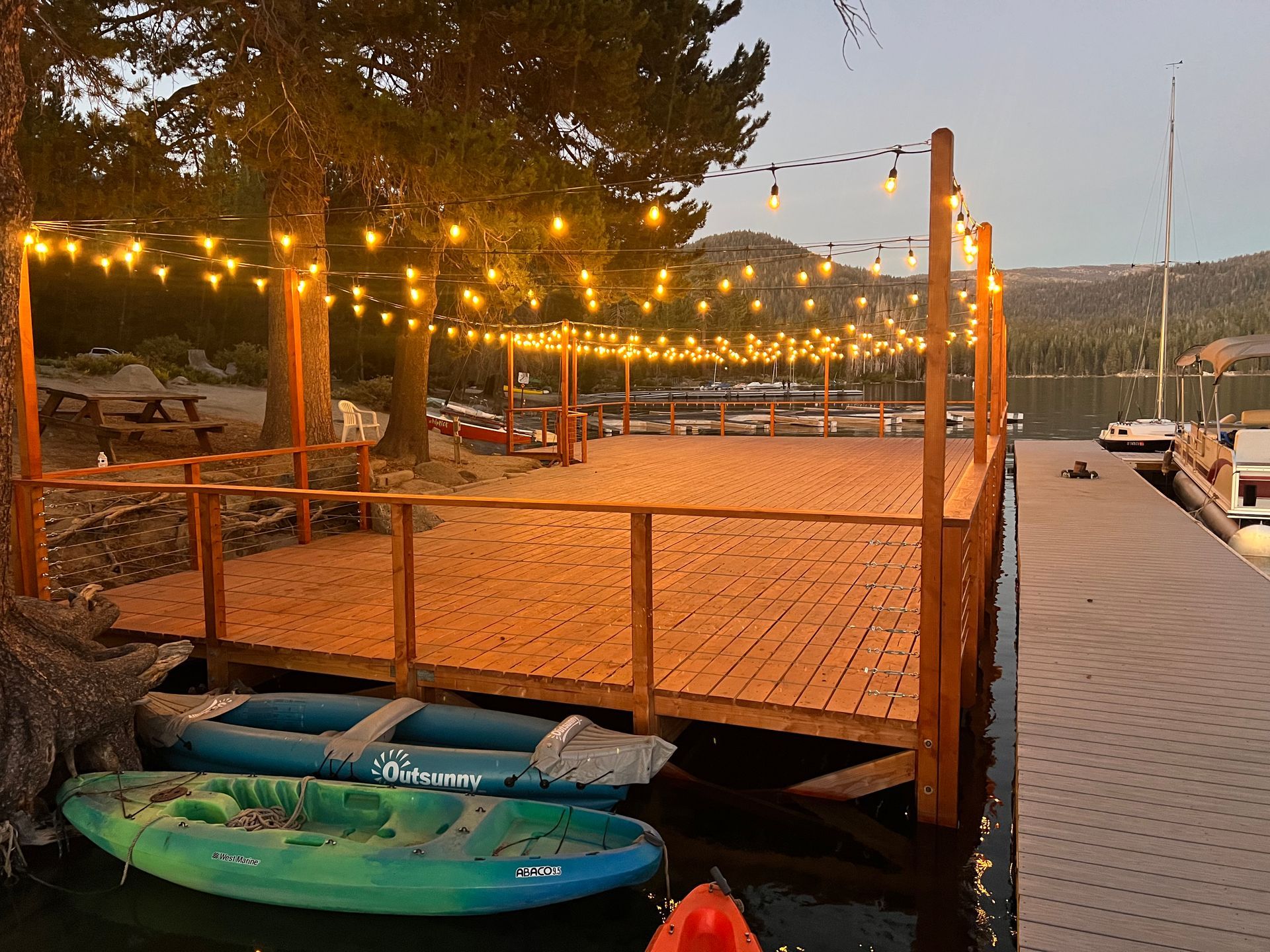 Wooden dock with string lights at sunset, a green kayak below, and chairs on a lakeside deck.