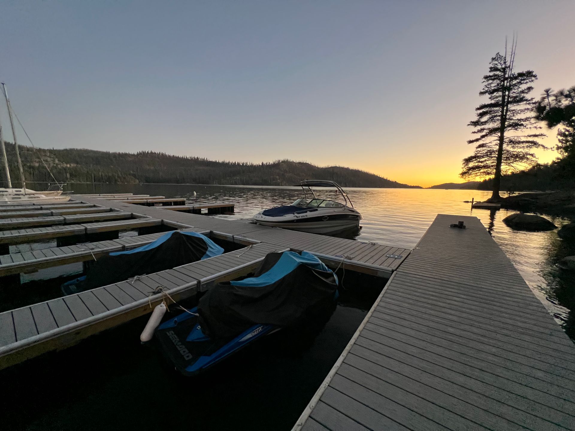 Snowy dock by a calm lake at sunset, with moored boats and silhouetted trees.
