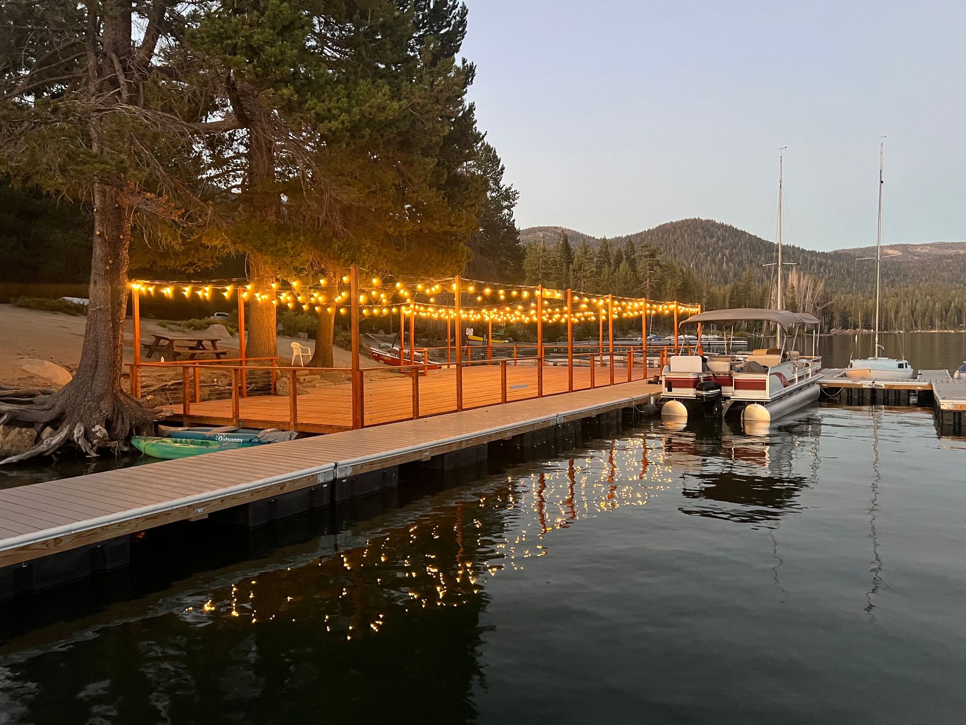 Docks at dusk with warm string lights, trees, and small boats on calm water by a marina