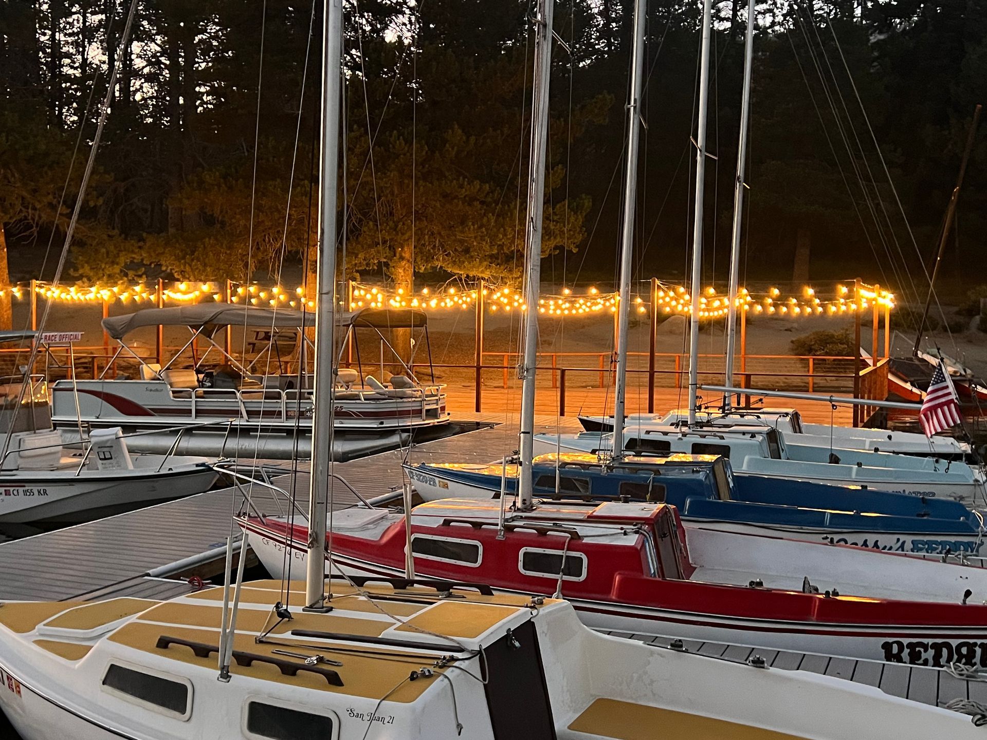 Sailboats docked at dusk with lit shoreline and reflections on calm water.