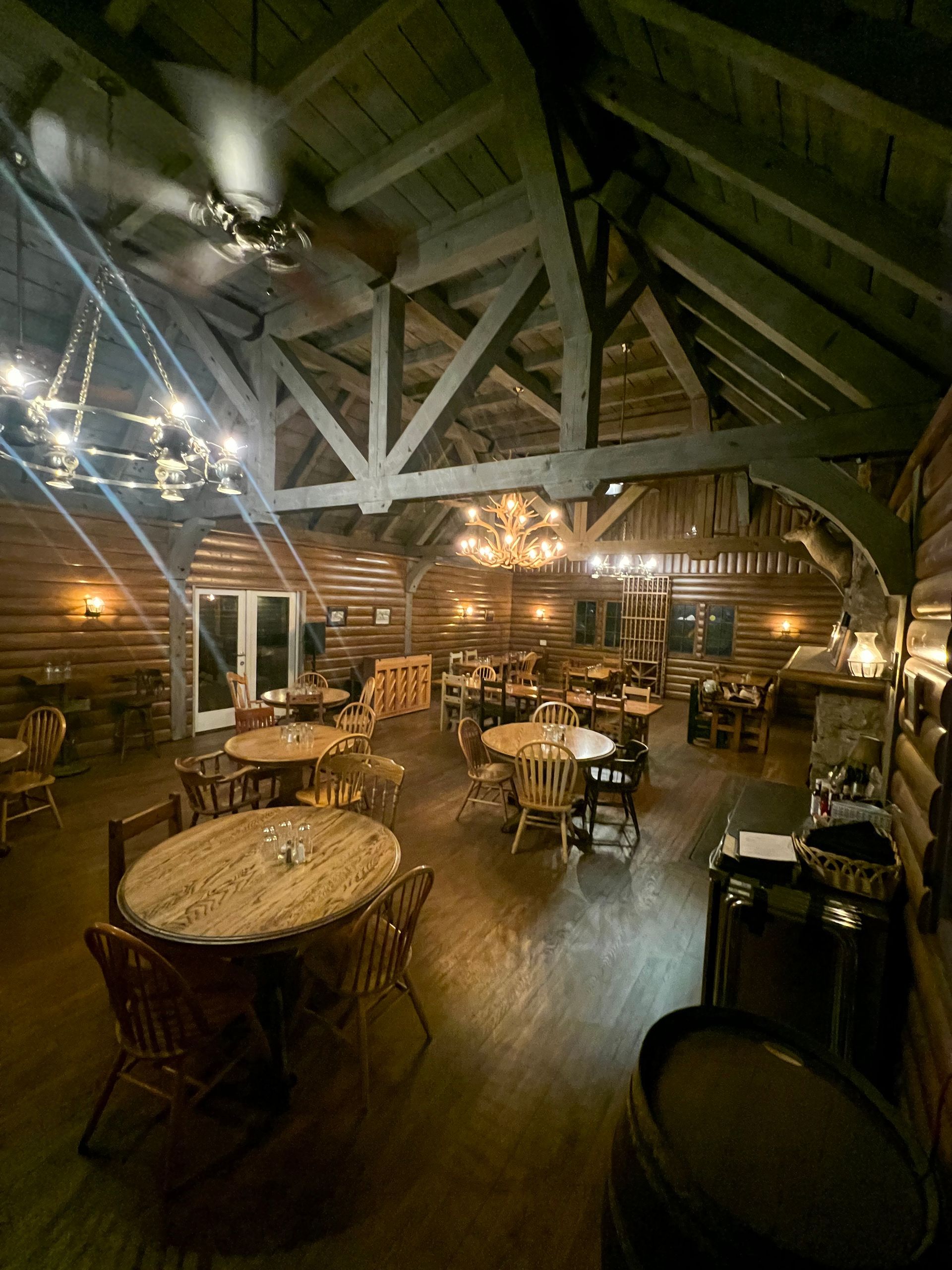 Rustic indoor dining hall with wooden tables, chairs, and warm string lights under exposed beams