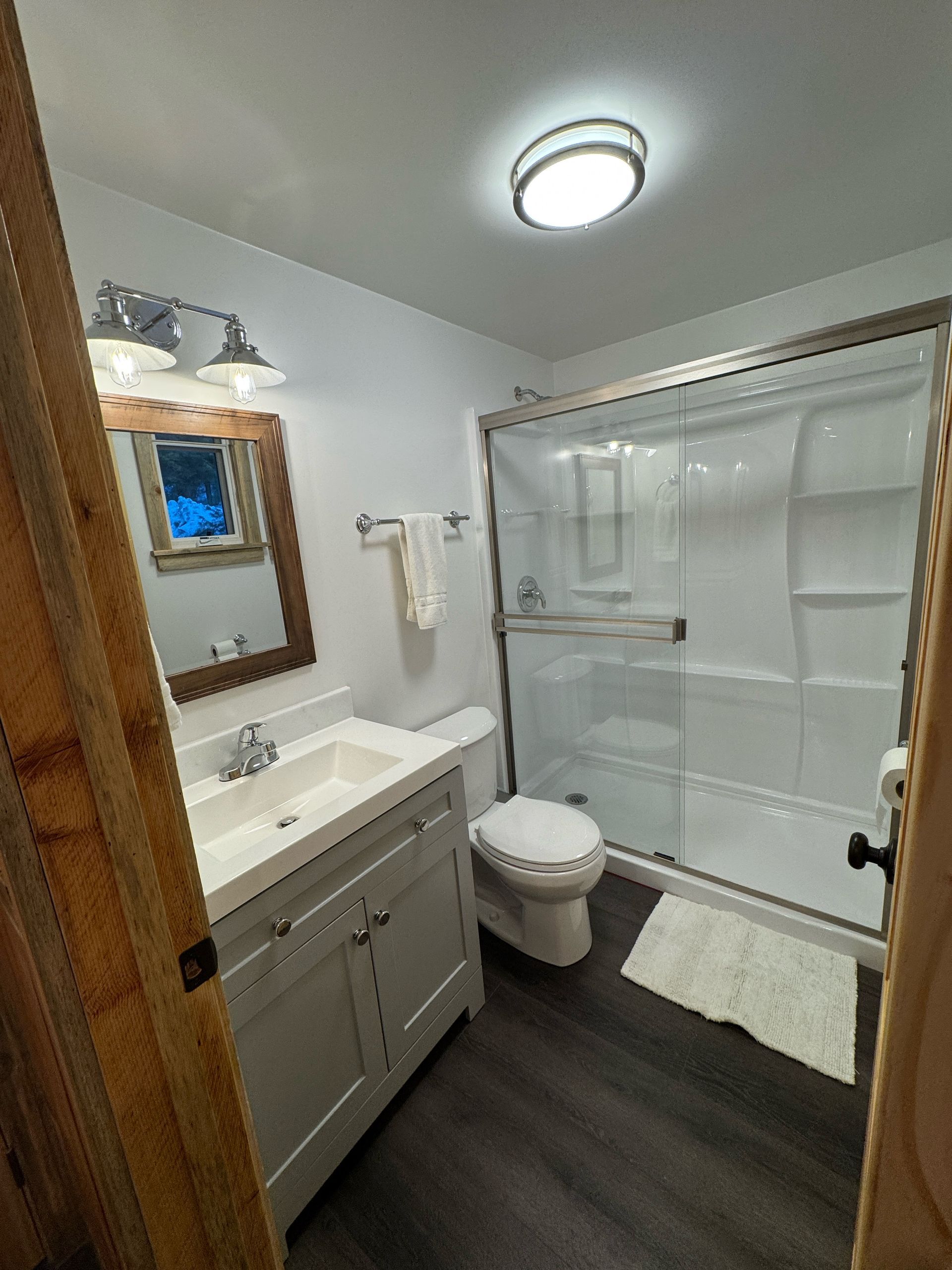 Small bathroom with white vanity, toilet, glass shower, dark floor, and a windowed mirror.
