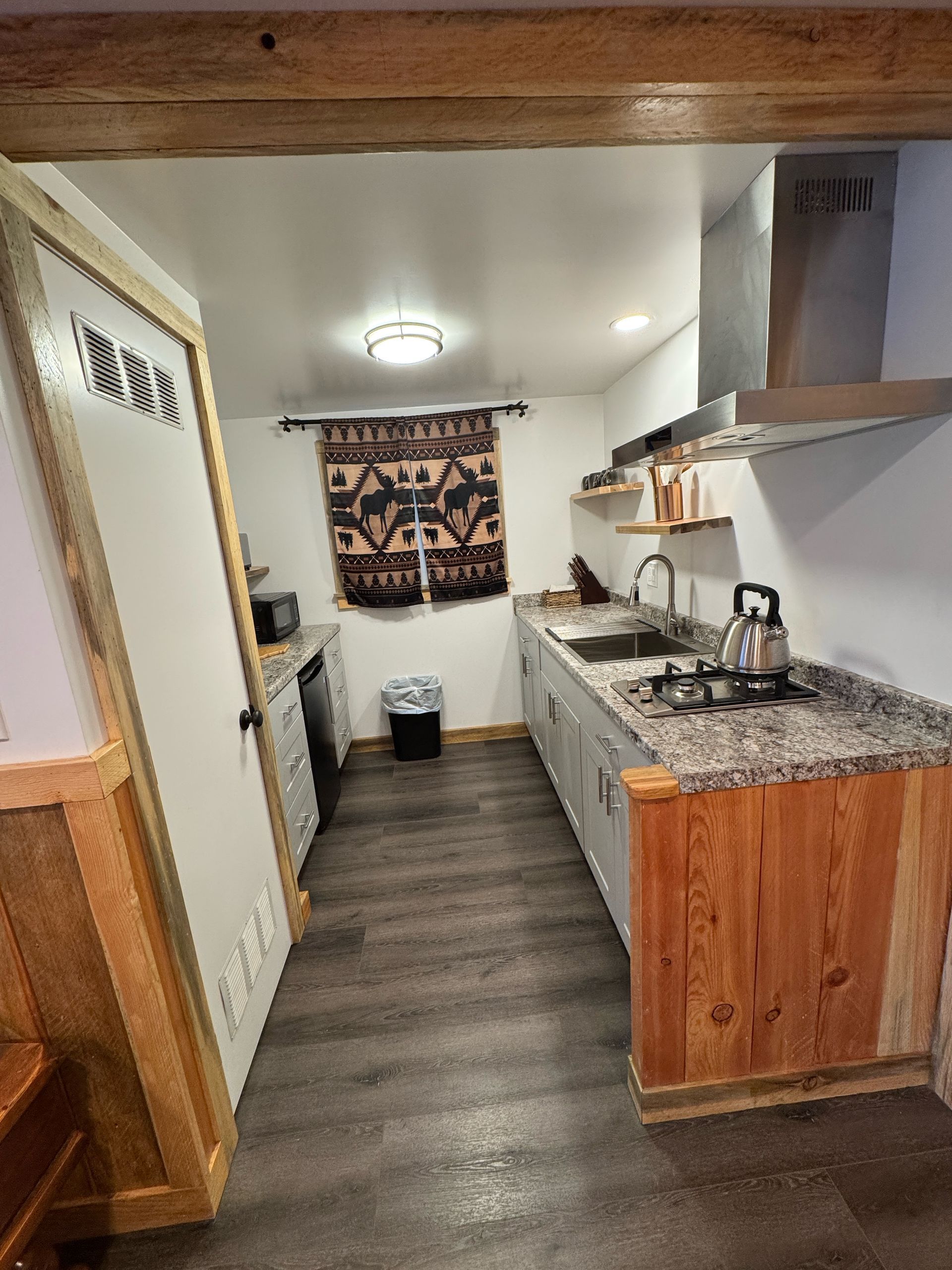 Narrow kitchen with wood cabinets, granite countertop, stove hood, and a small window above a trash can.