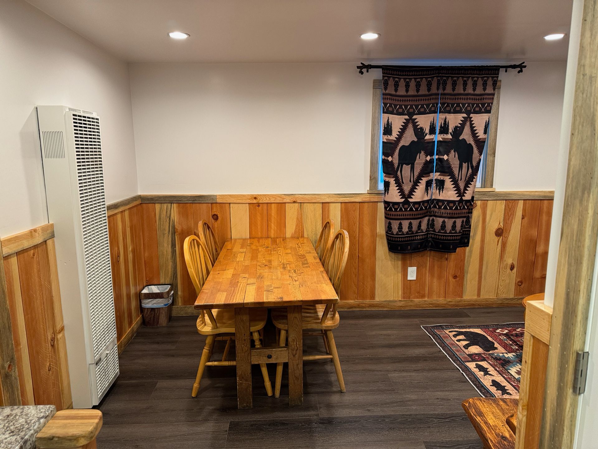 Dining room with wooden table and chairs, paneled walls, dark floor, and a hanging tapestry.