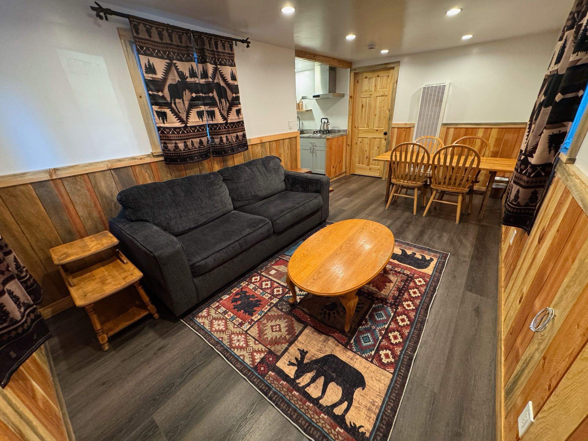 Cozy living room with black sofa, wooden coffee table, patterned rug, and dining area in the background
