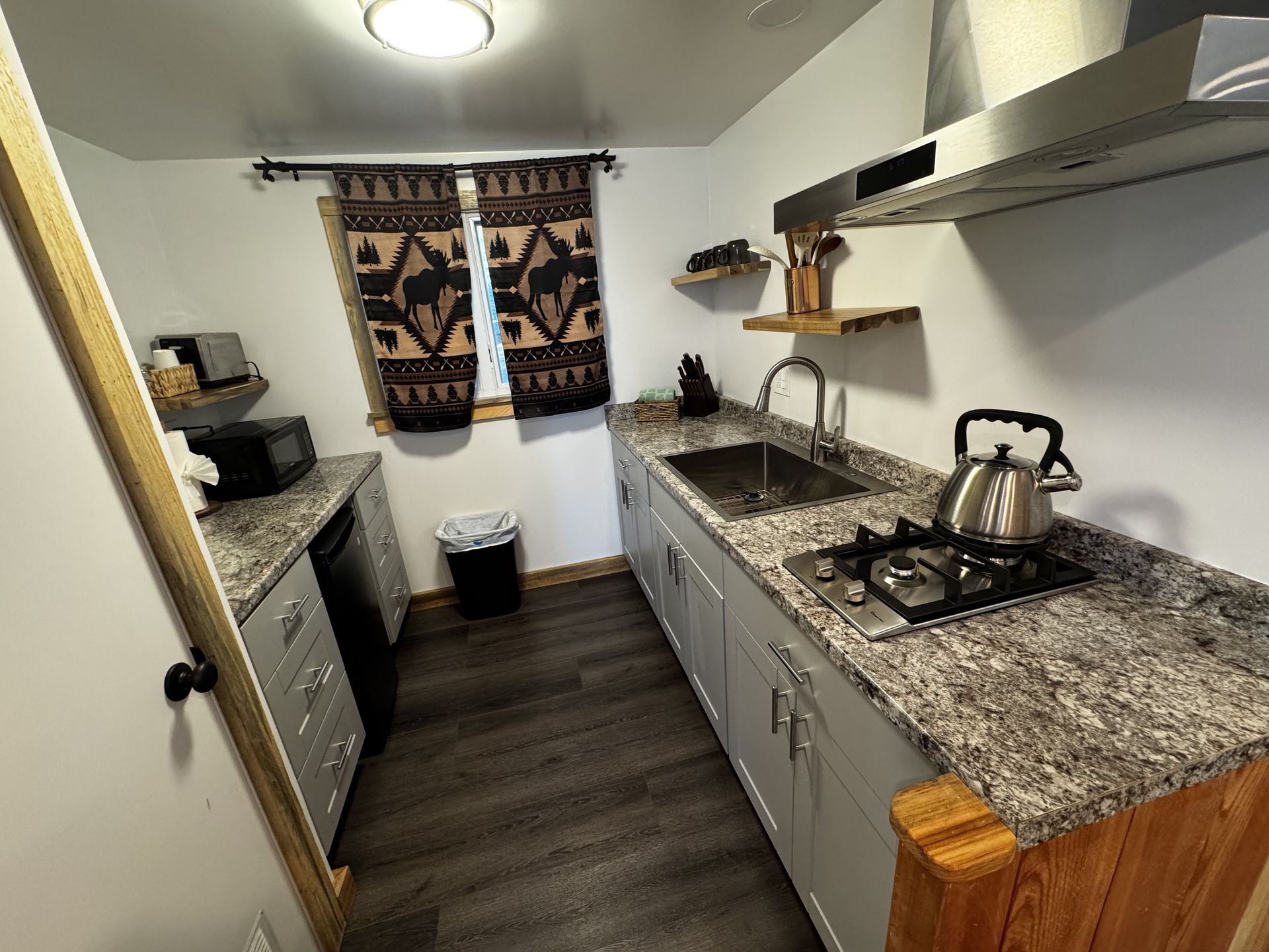 Narrow galley kitchen with granite countertops, stainless stove and kettle, white cabinets, and patterned curtains.