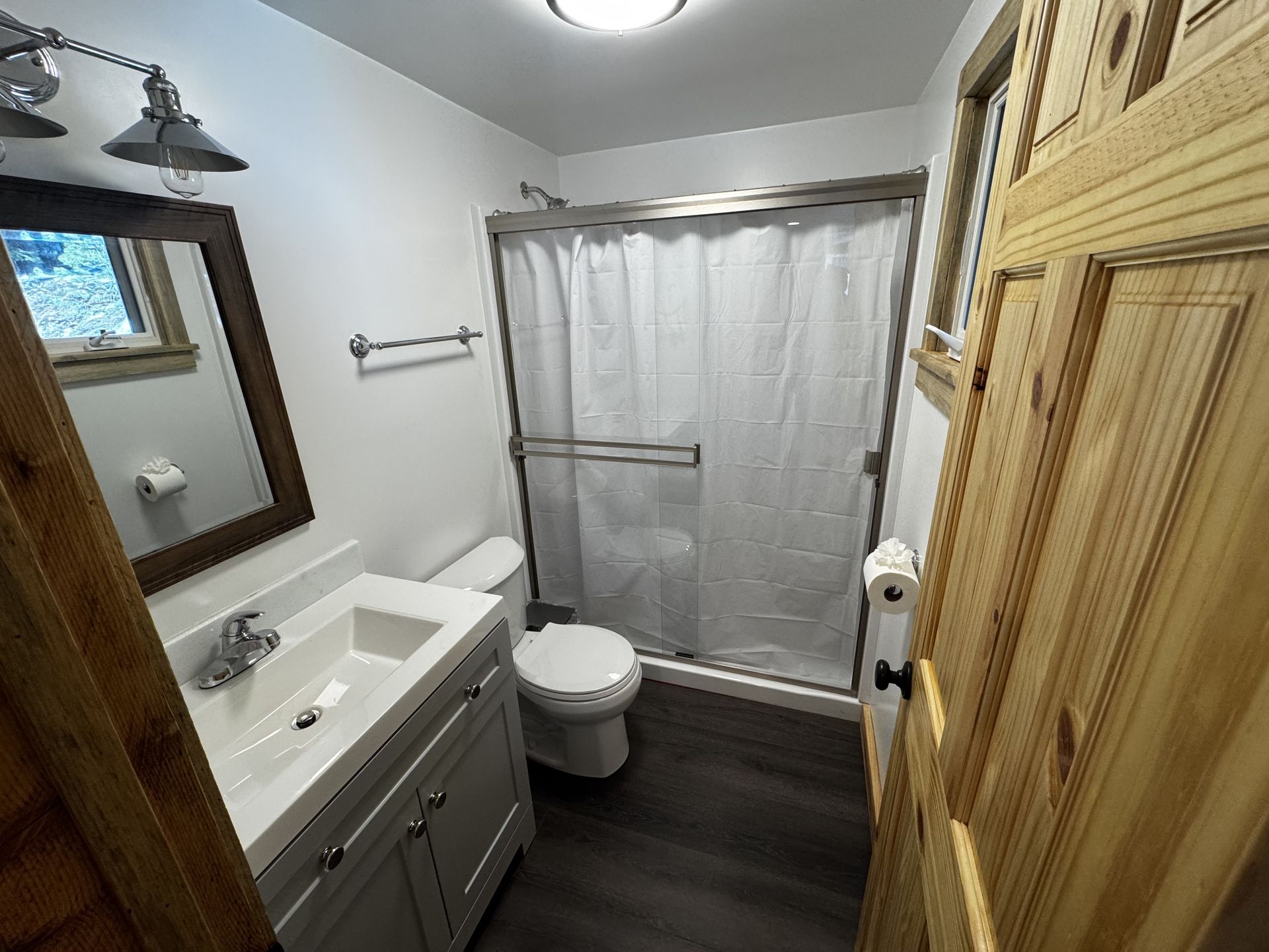 Small bathroom with gray vanity, toilet, shower curtain, wood floor, and wall mirror.