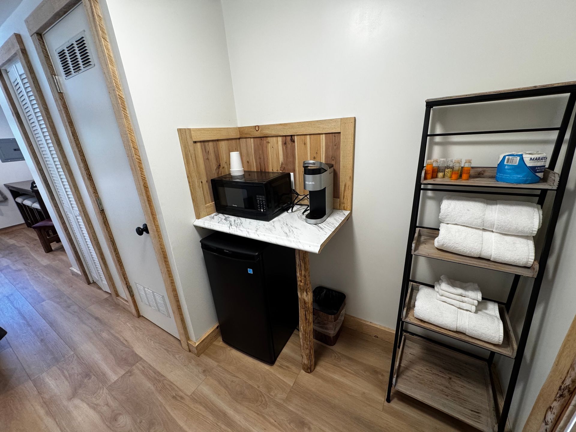 Bathroom corner with black mini fridge, coffee station, and metal shelf holding towels and toiletries