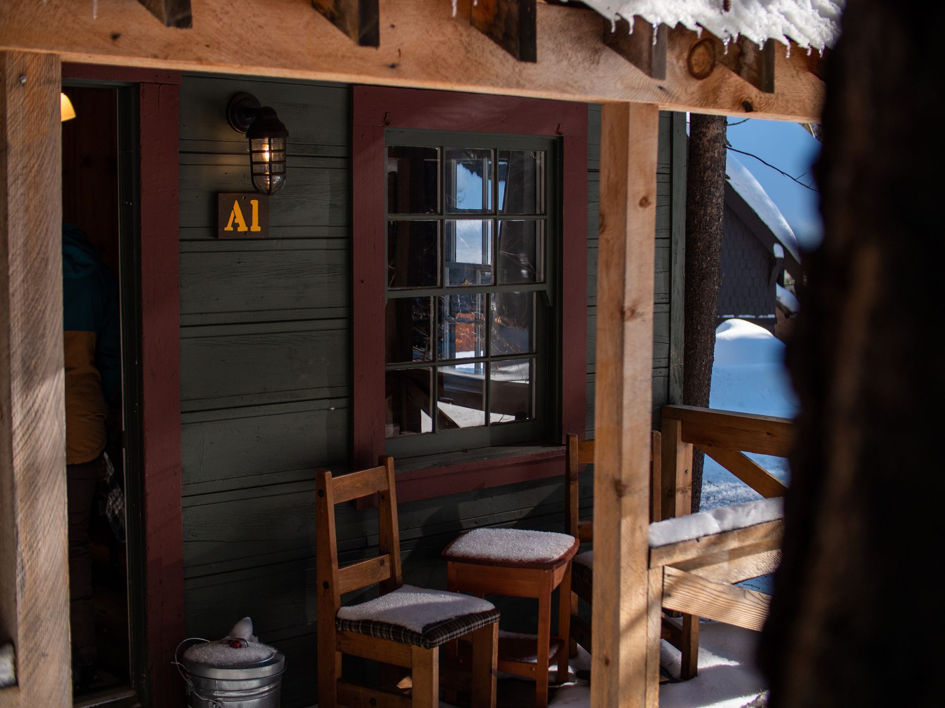 Cozy rustic porch with chairs, table, and lantern beside a wooden cabin door in snowy weather
