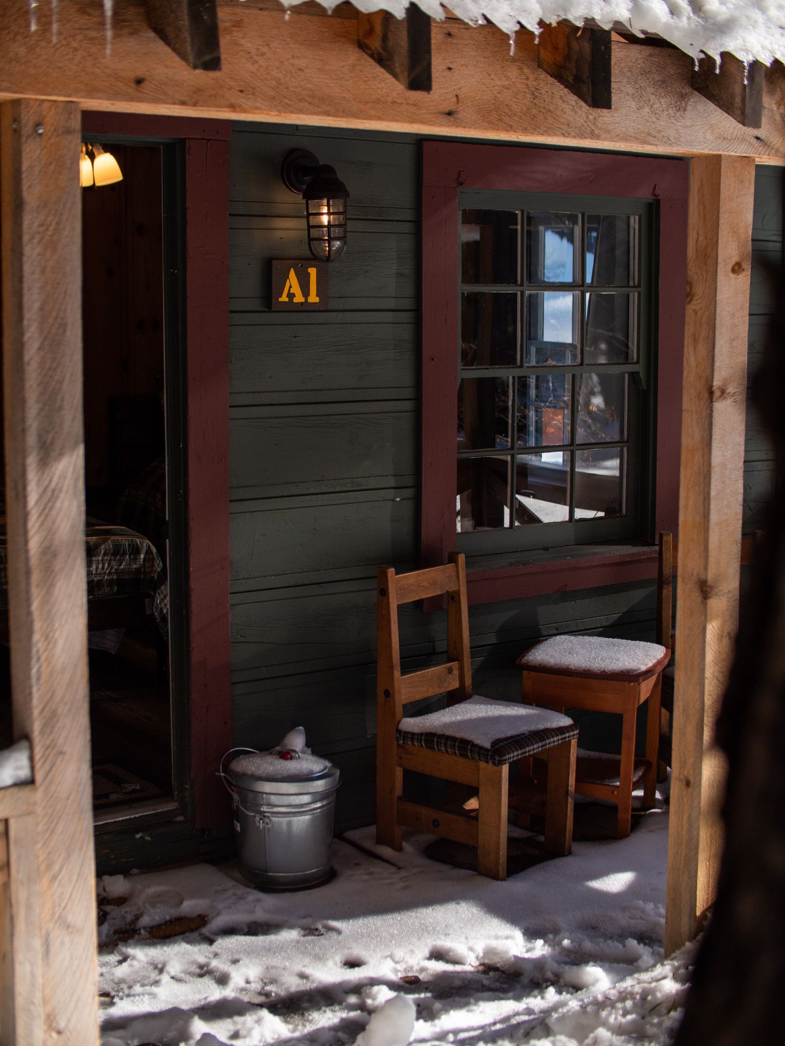 Snowy rustic porch with a wooden chair, bench, and a dark door marked “A1” beside a window