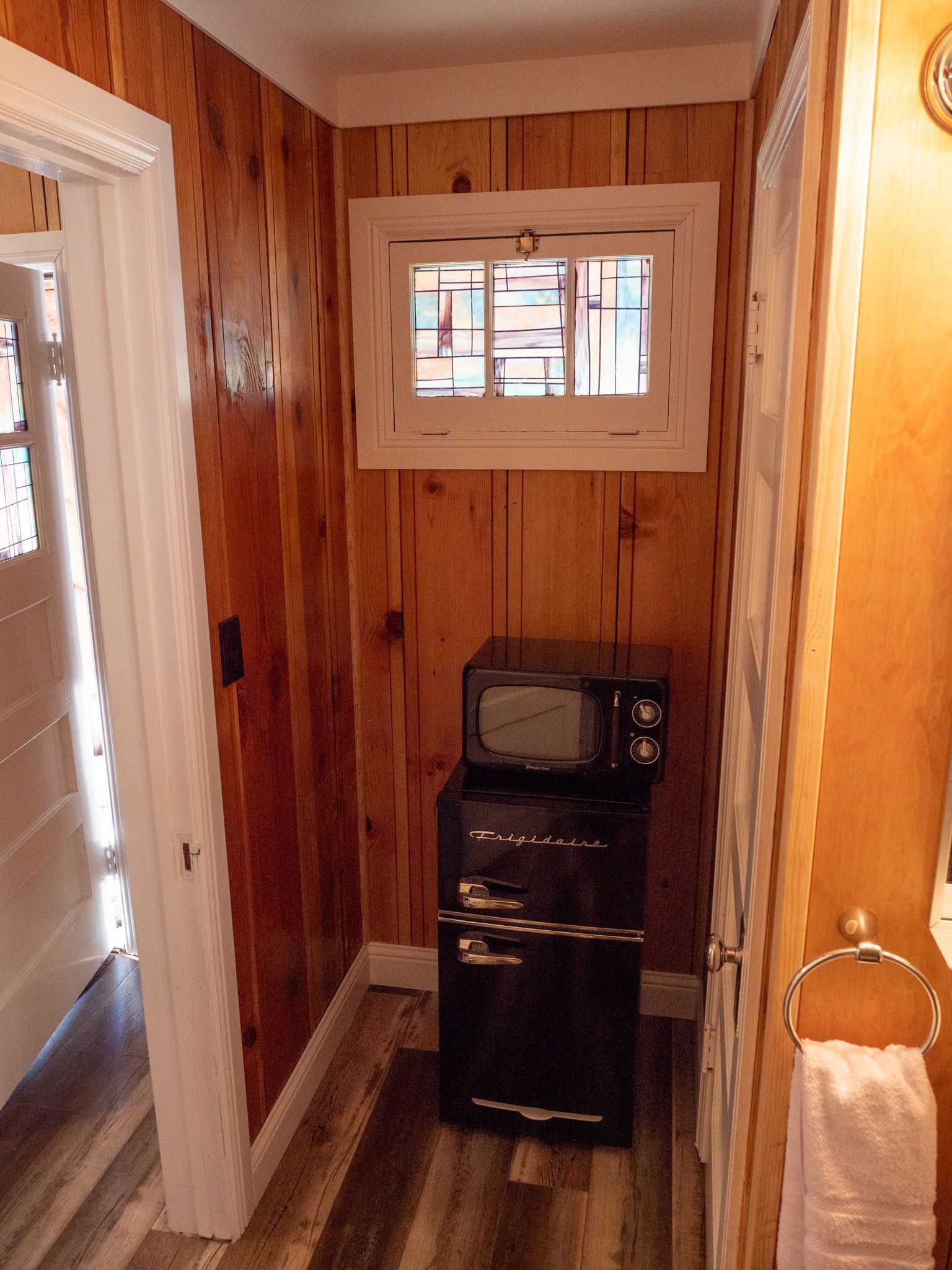 Narrow wood-paneled hallway with a small window, mini fridge, microwave, and towel ring.