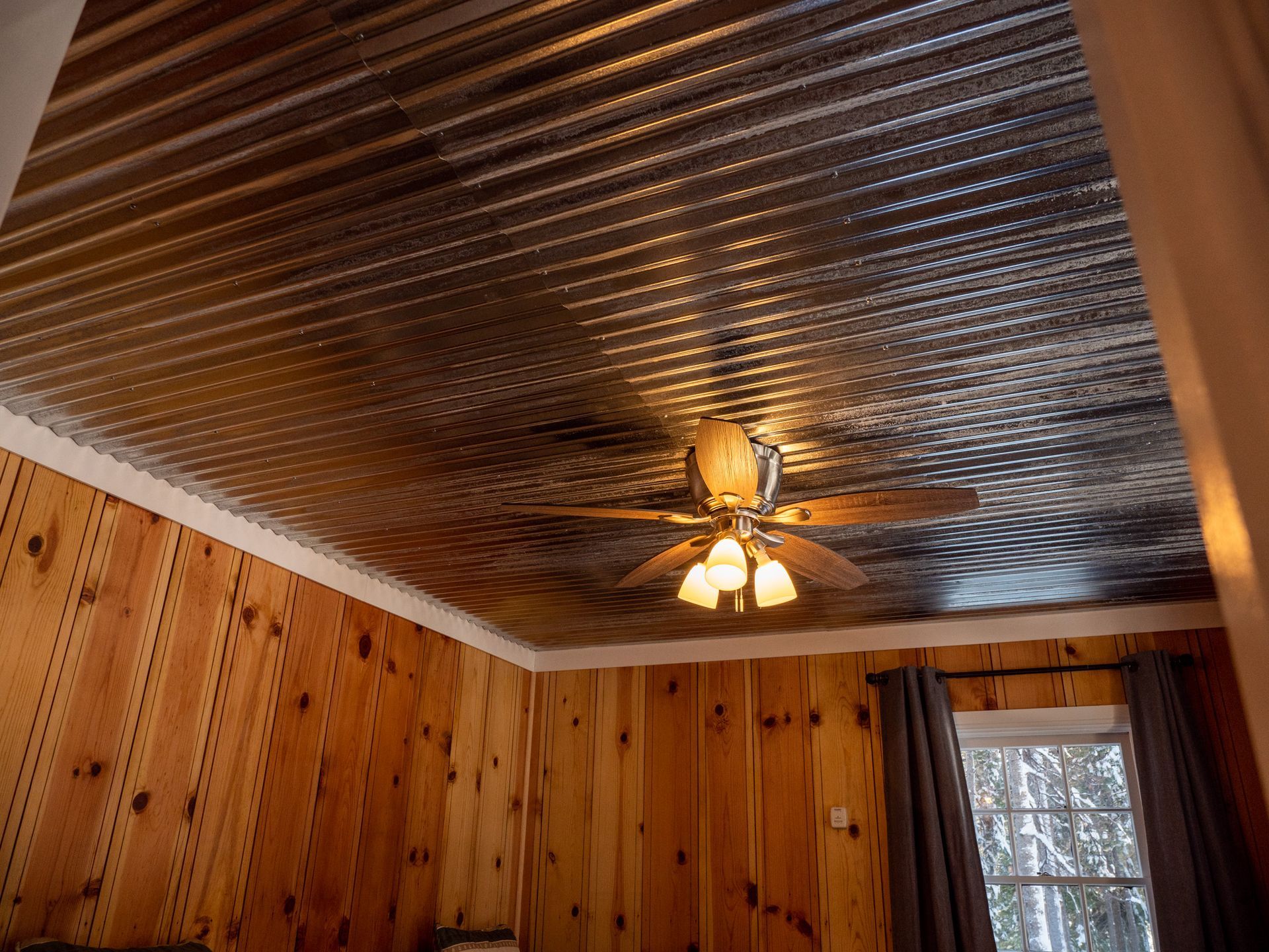 Wood-paneled room with a corrugated metal ceiling, ceiling fan, and a window with curtains.