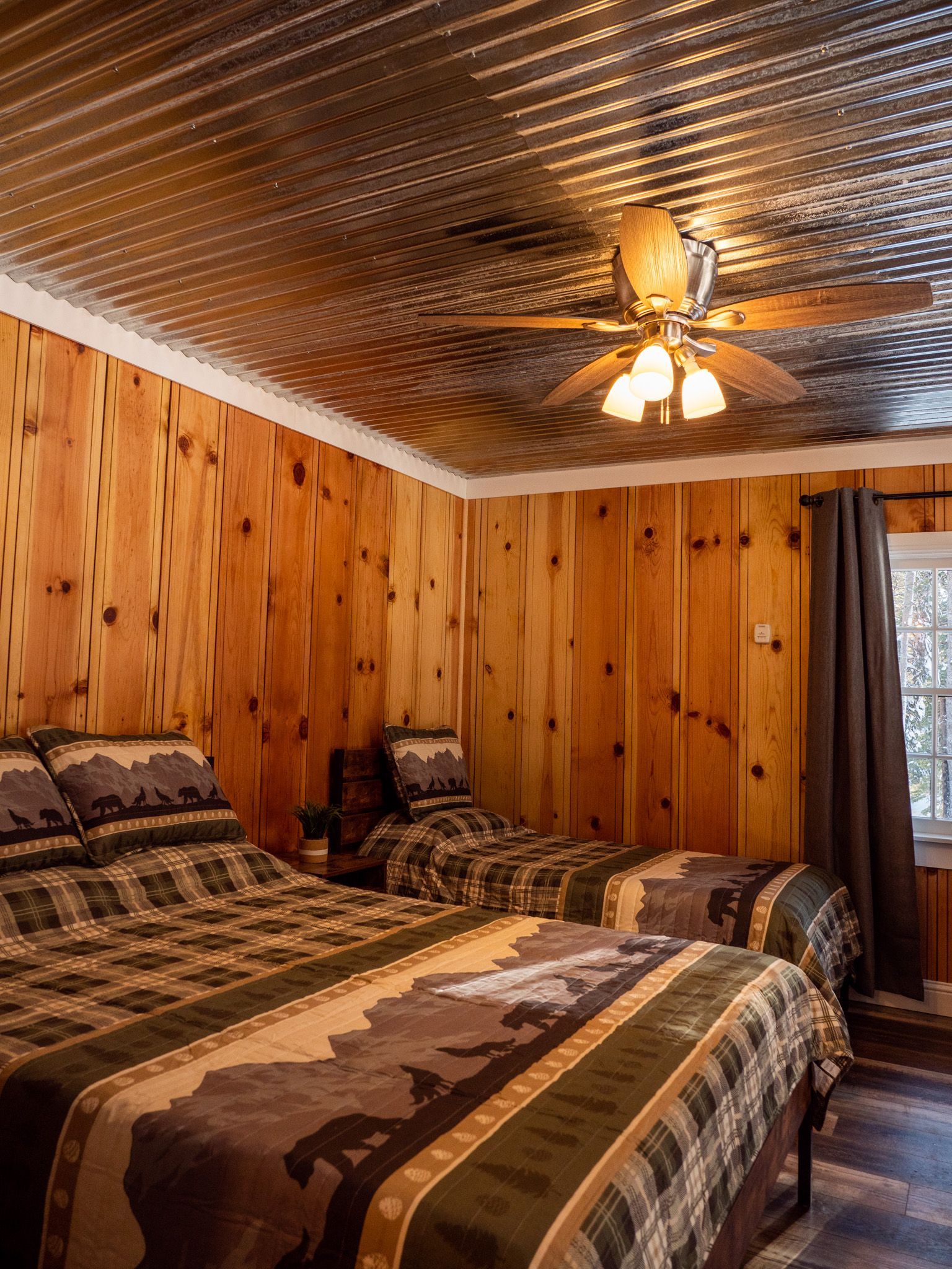 Cozy bedroom with two patterned beds, wood-paneled walls, metallic ceiling, and a ceiling fan.