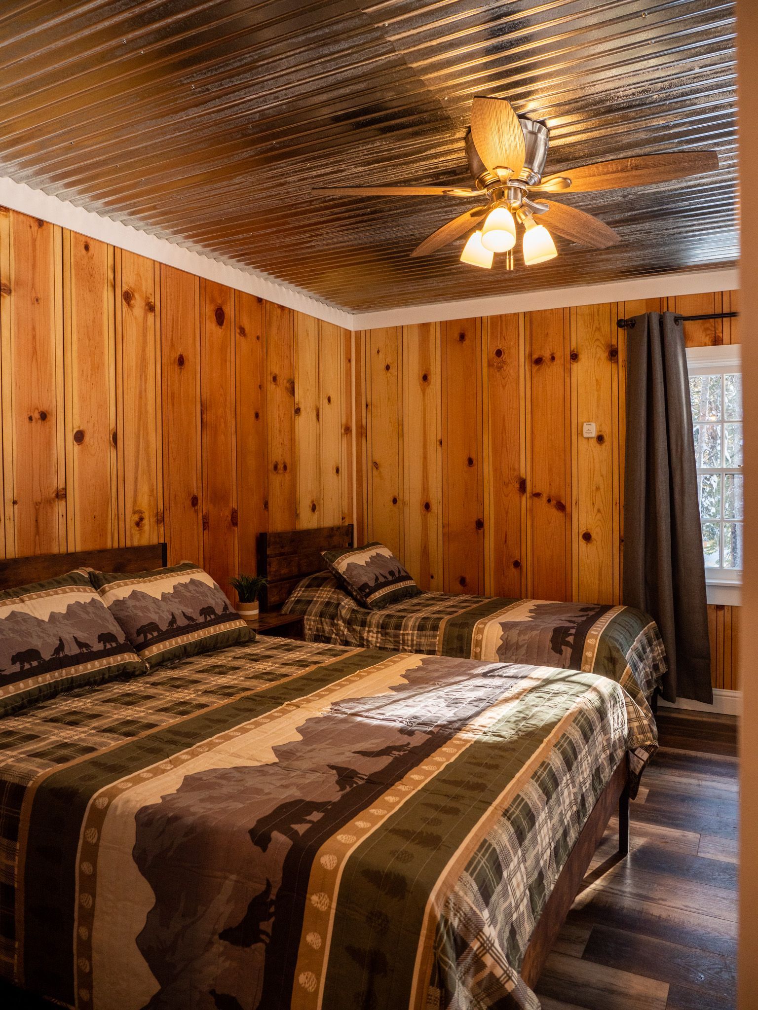 Rustic bedroom with wood-paneled walls, two beds, patterned bedding, and a ceiling fan with lights