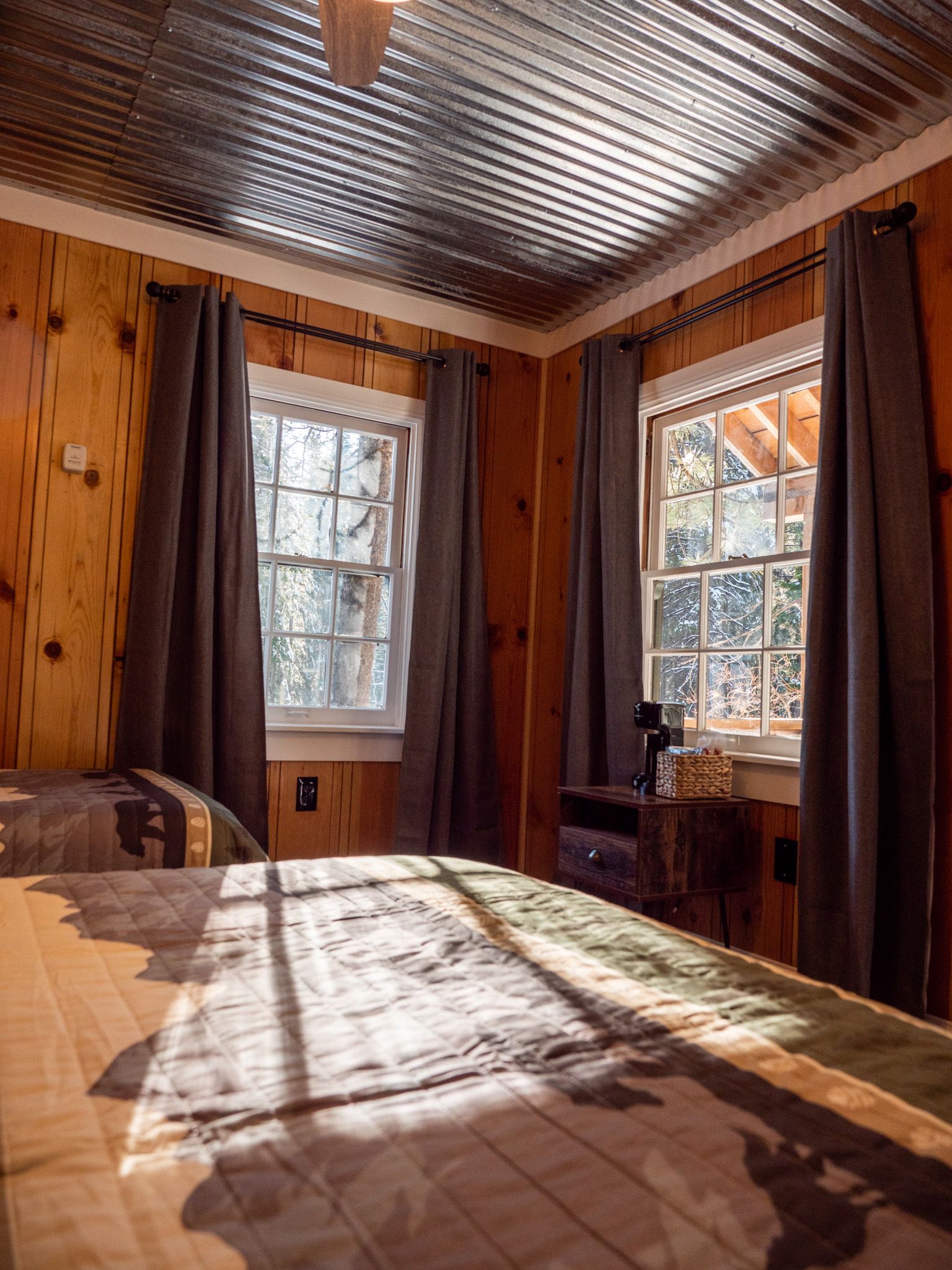 Sunlit rustic bedroom with wood-paneled walls, two windows, and a bed in the foreground