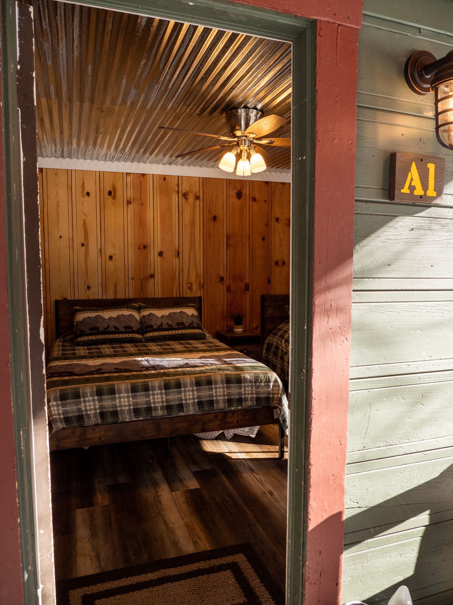 Cozy rustic bedroom with wood-paneled walls, plaid bedspread, and a ceiling fan in a narrow doorway view