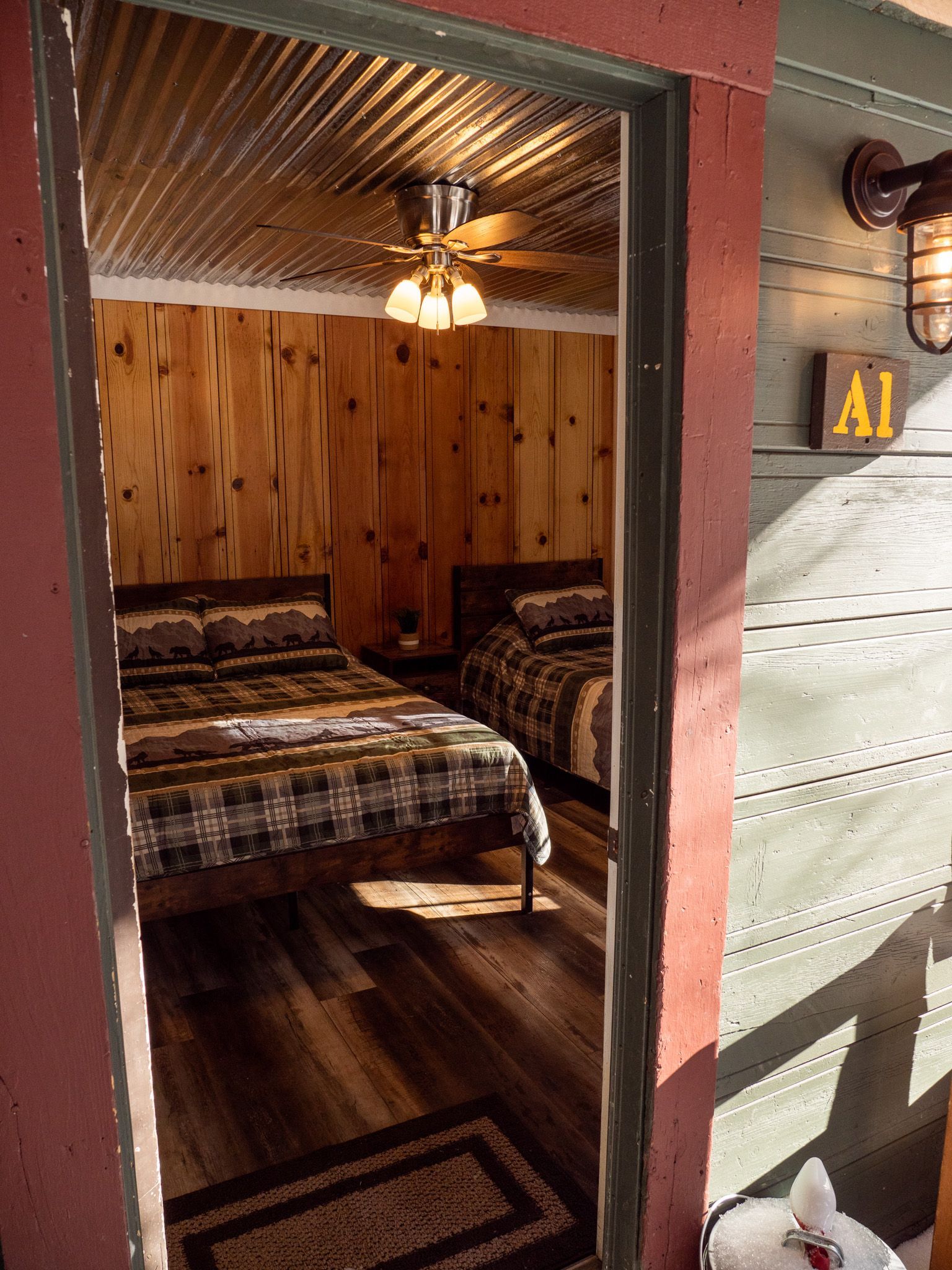 Cozy wooden room with two beds, ceiling fan, and patterned rug, viewed from an open doorway.