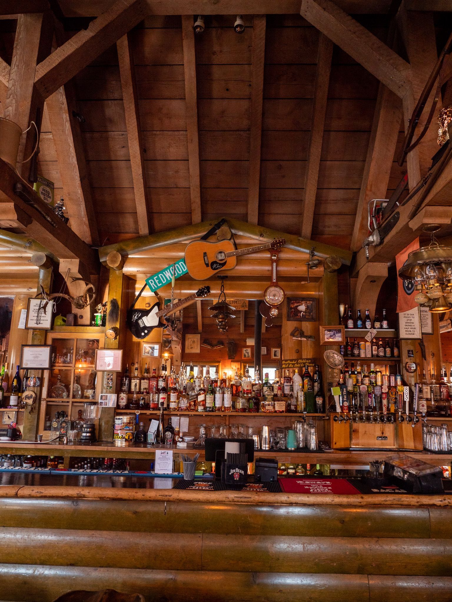 Rustic wooden bar interior with shelves of bottles, mounted decor, and a log counter under exposed beams.