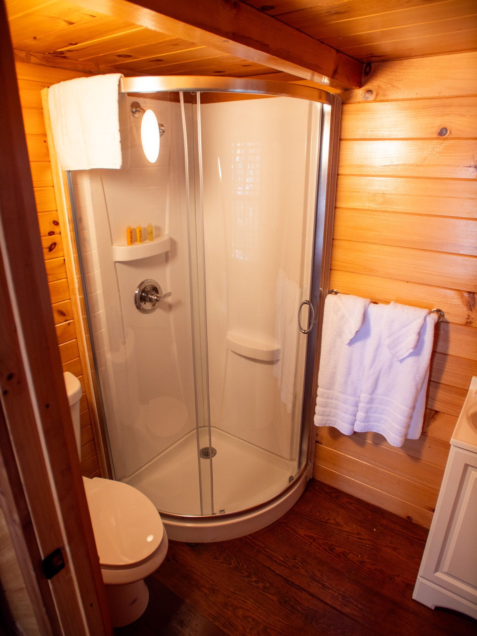 Small wooden bathroom with a corner shower, toilet, towel rack, and white cabinetry.