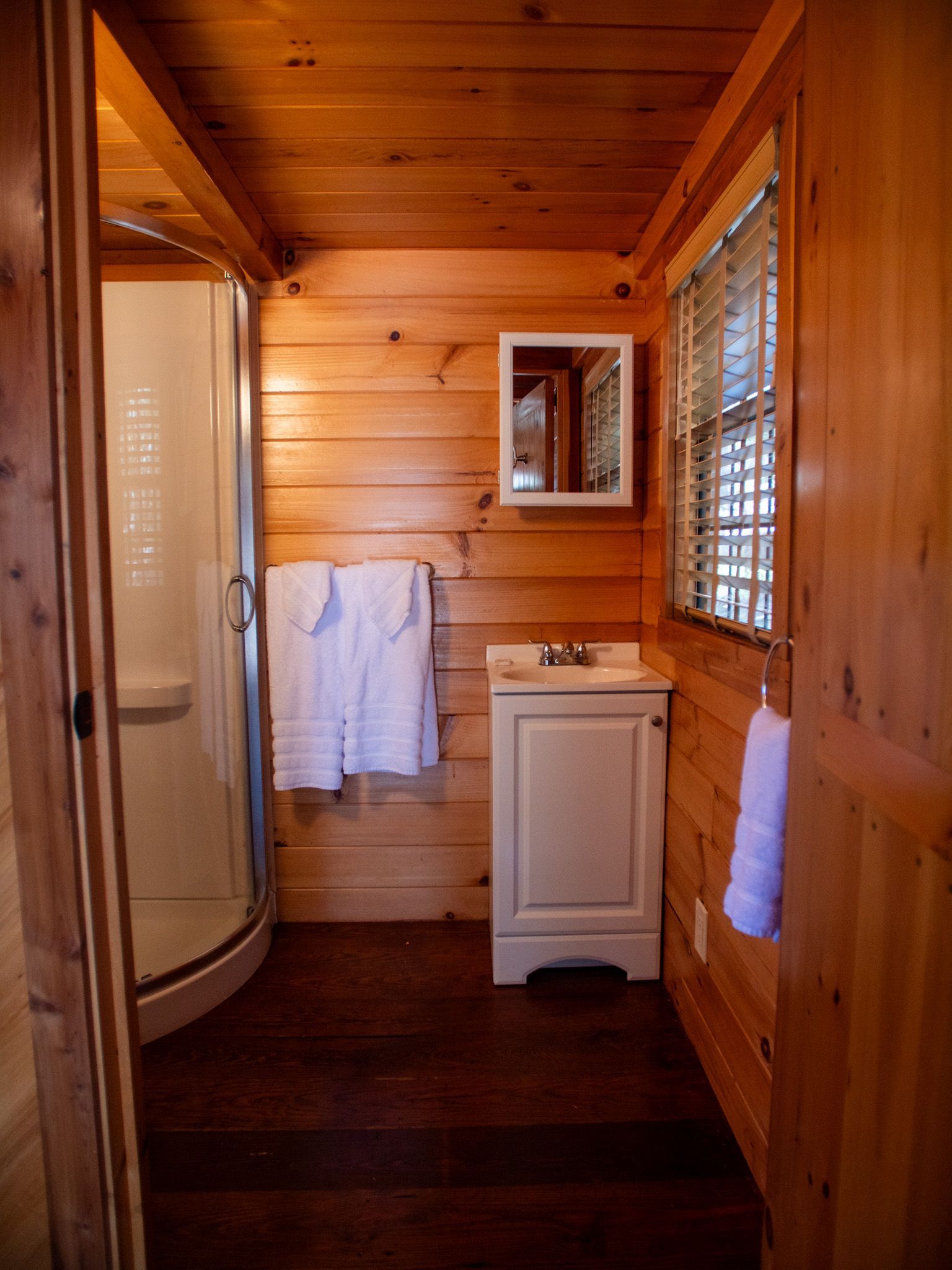 Wood-paneled bathroom with shower, sink, mirror, and white towels by a window and cabinet.