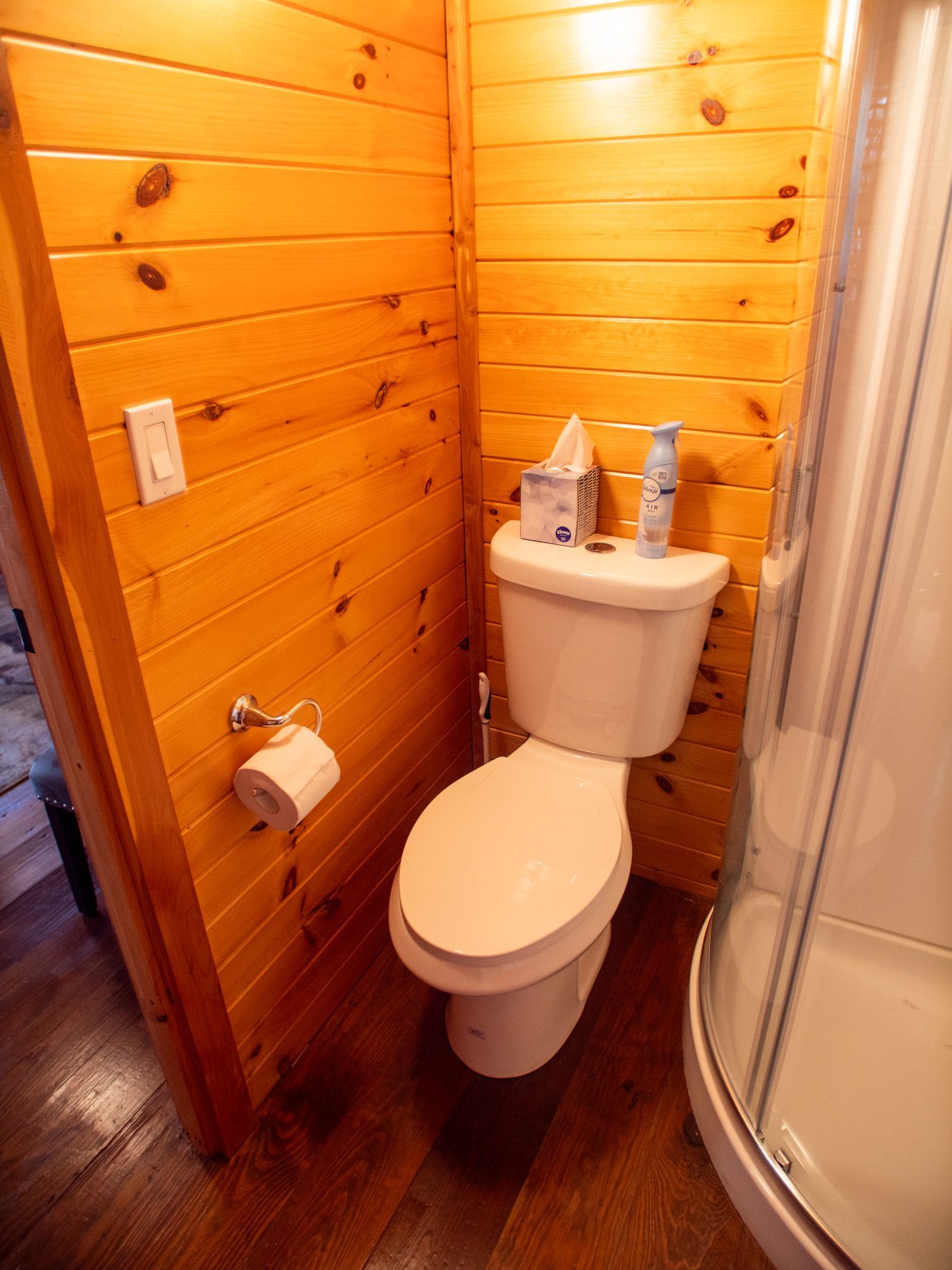 Small wooden bathroom with a toilet, sink, toilet paper holder, and shower stall.