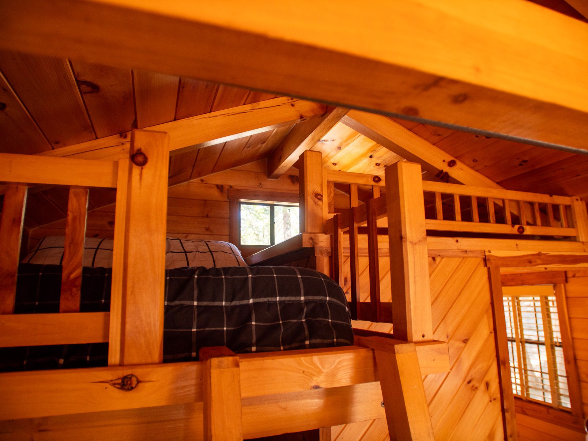 Wooden loft interior with exposed beams, a bed, and a small window through to a room below.