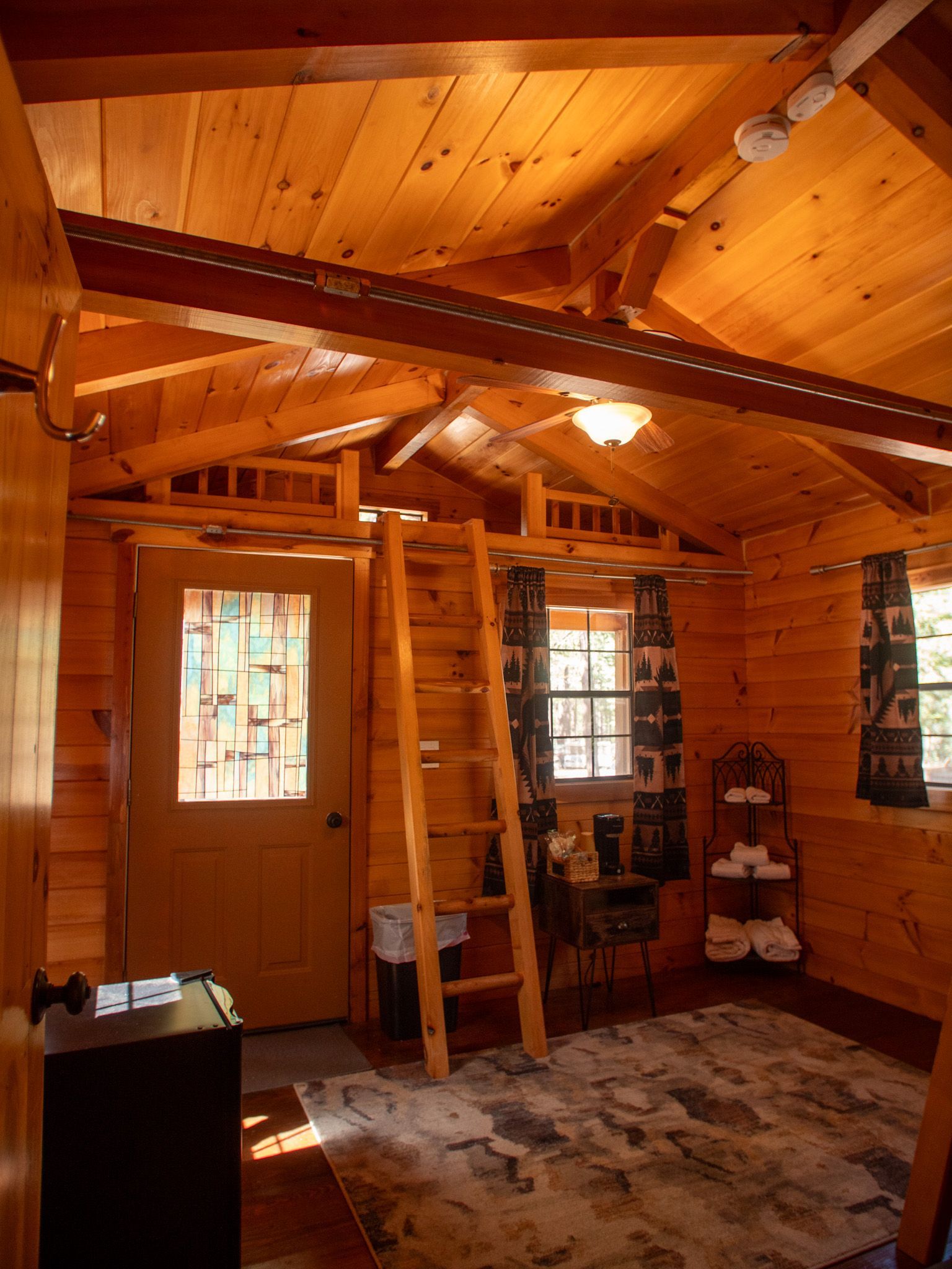 Wooden cabin interior with exposed beams, ladder, windows, rug, and a door with sunlight streaming in