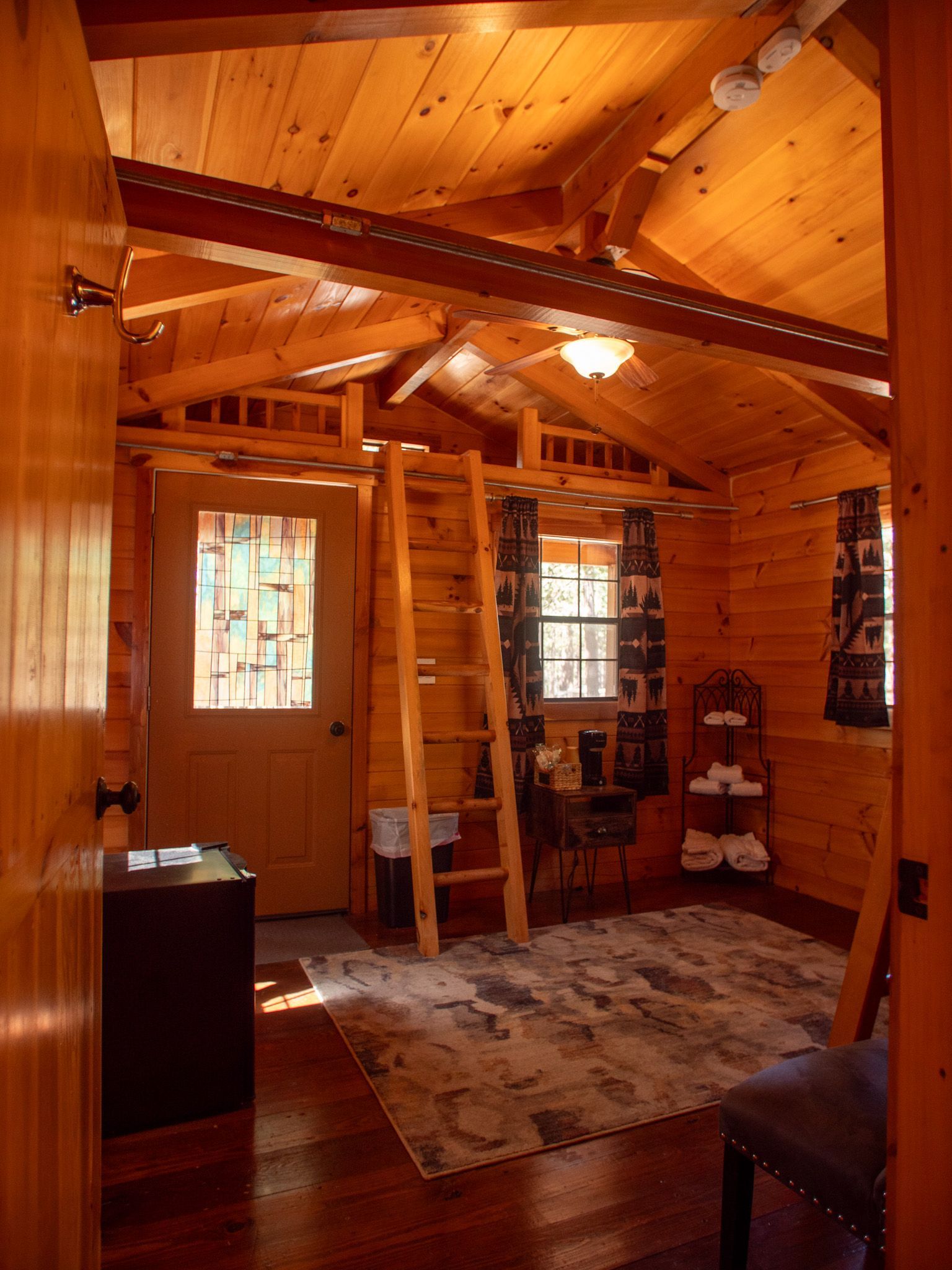 Warm wooden cabin interior with exposed beams, windows, rug, and a small chair