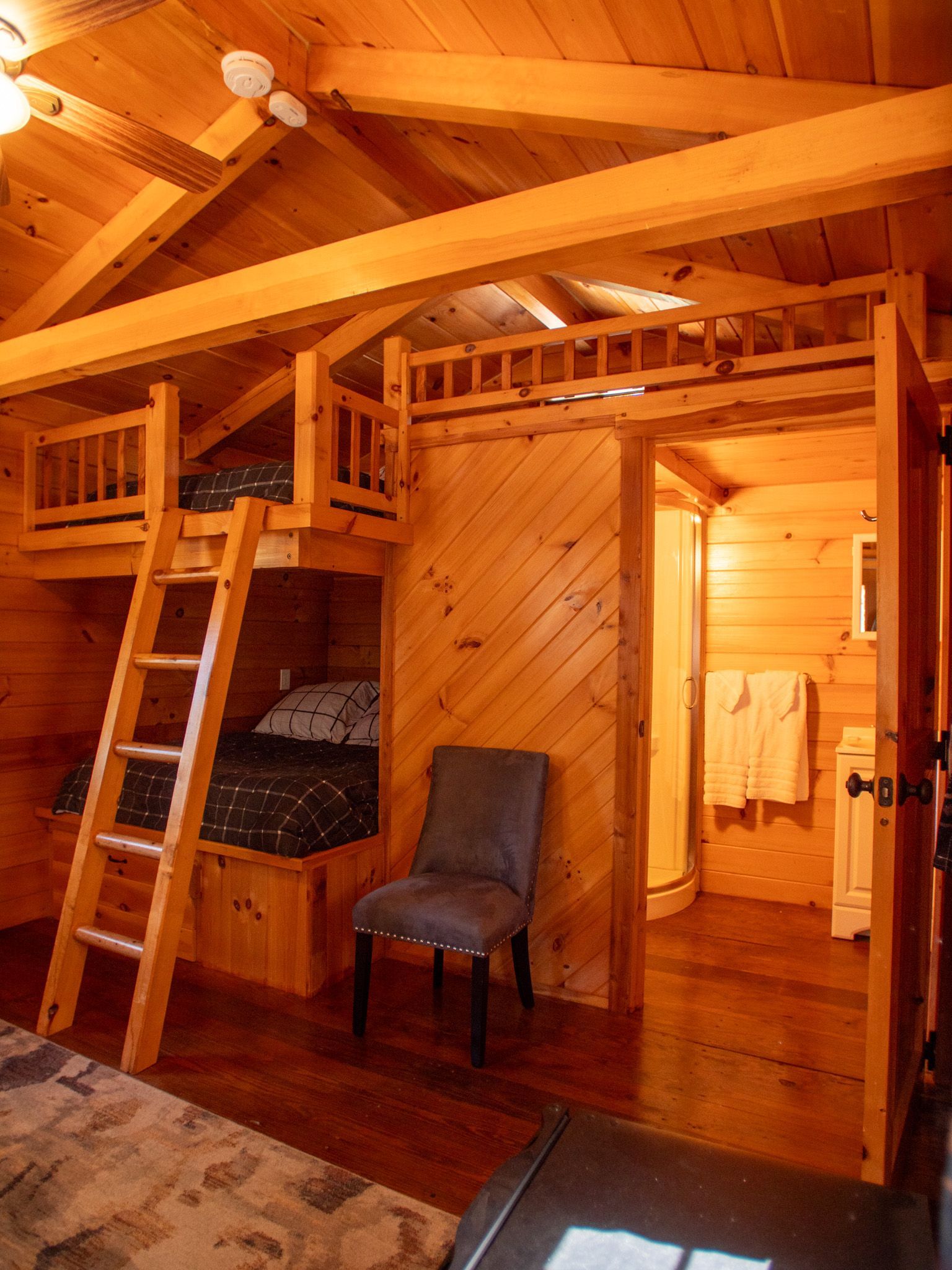 Warm wooden loft bedroom with bunk beds, ladder, chair, and open doorway in cabin interior