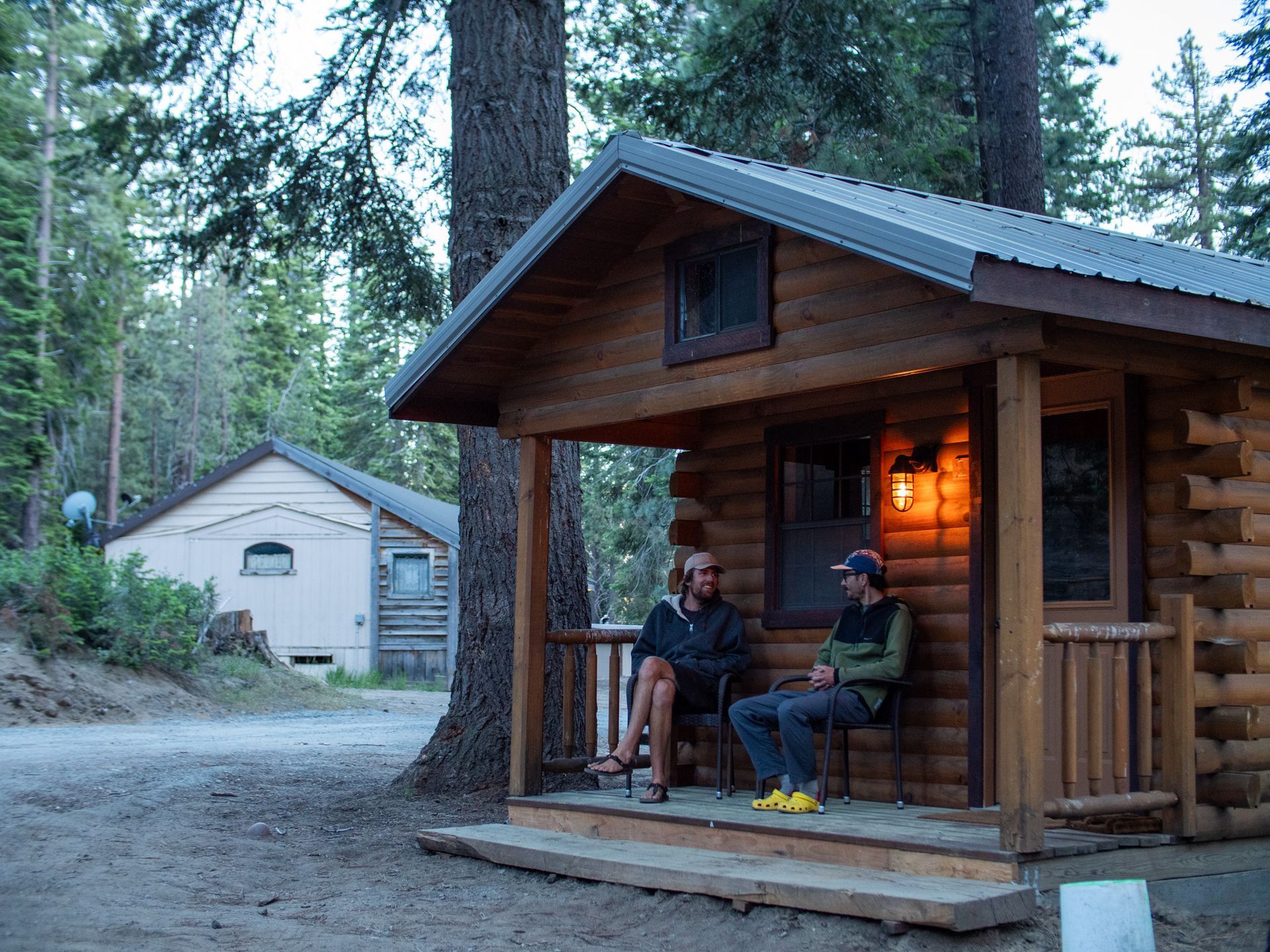 Two people sitting on a cabin porch at dusk, with warm porch lights and a small shed in the wooded background.
