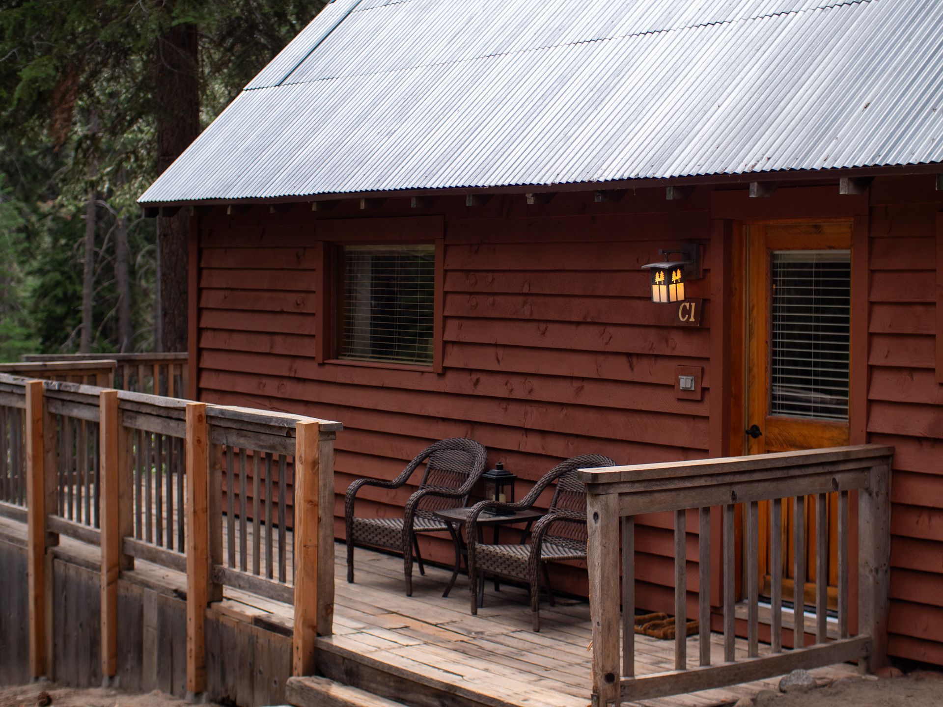 Wooden cabin porch with railing, bench, and lantern beside a window and door in the woods