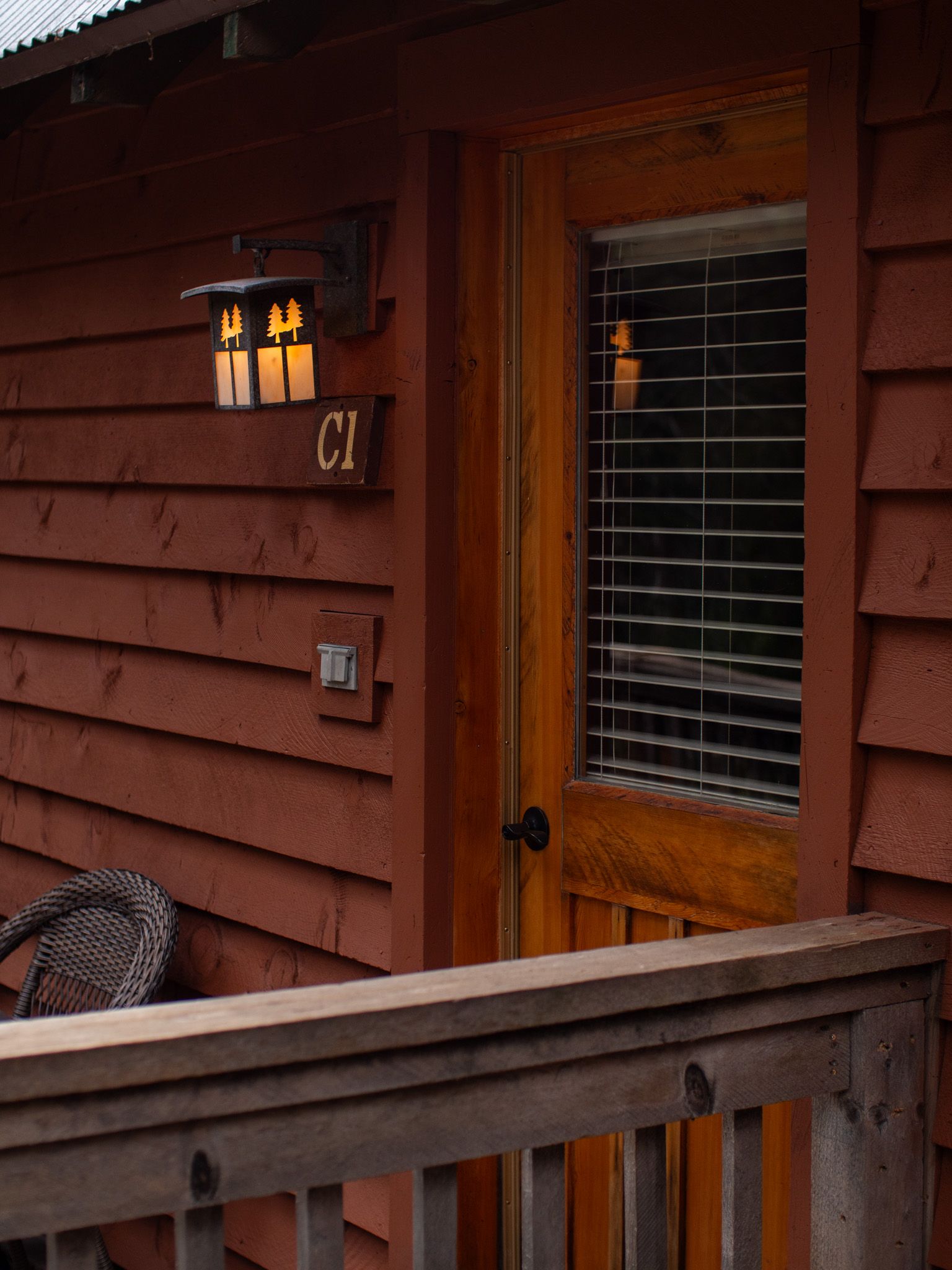 Wooden cabin porch with a lit wall lantern, window blinds, and railing in warm evening light
