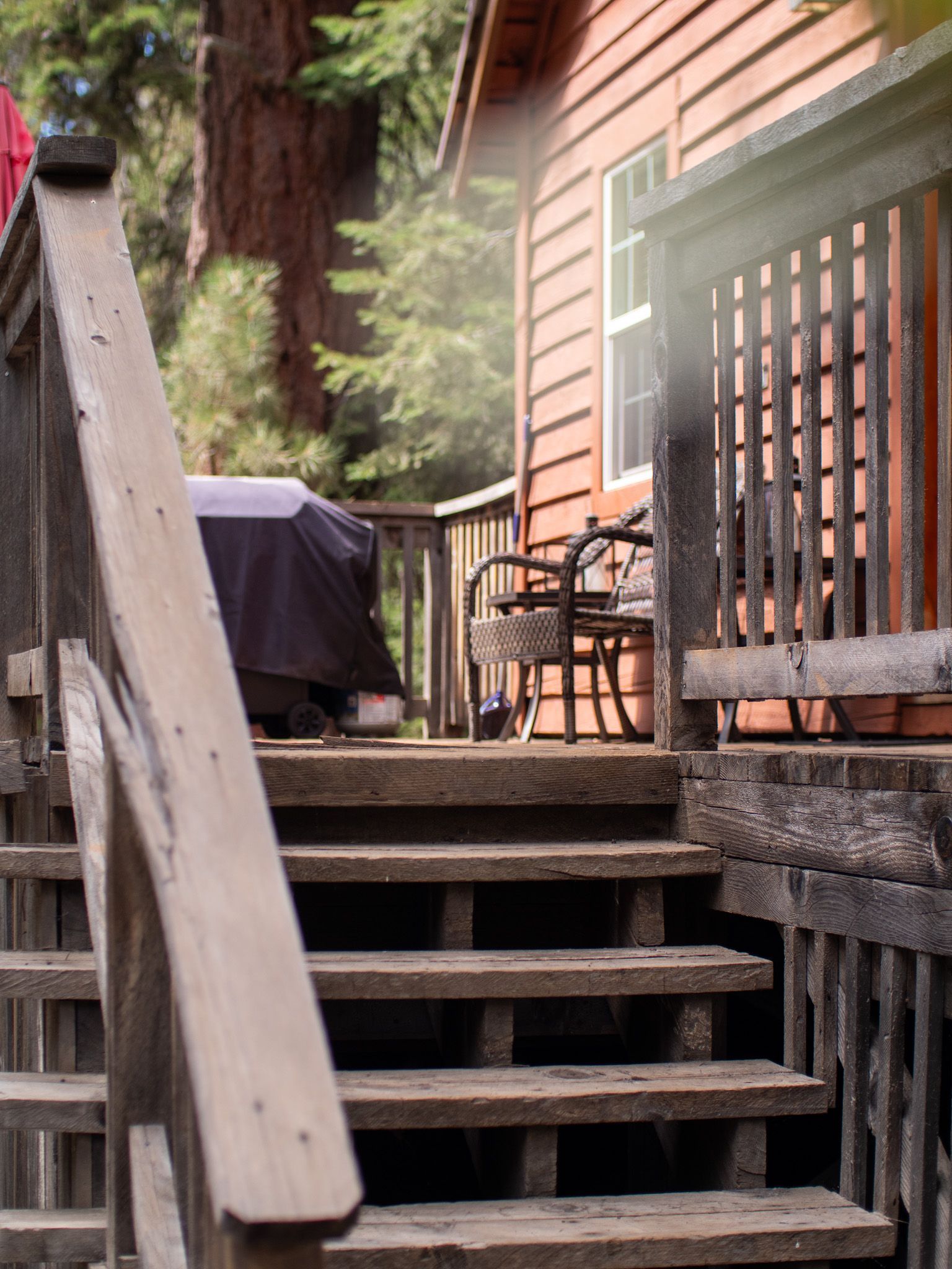 Wooden stairs beside a house deck with chairs, a grill, and tall trees in the background