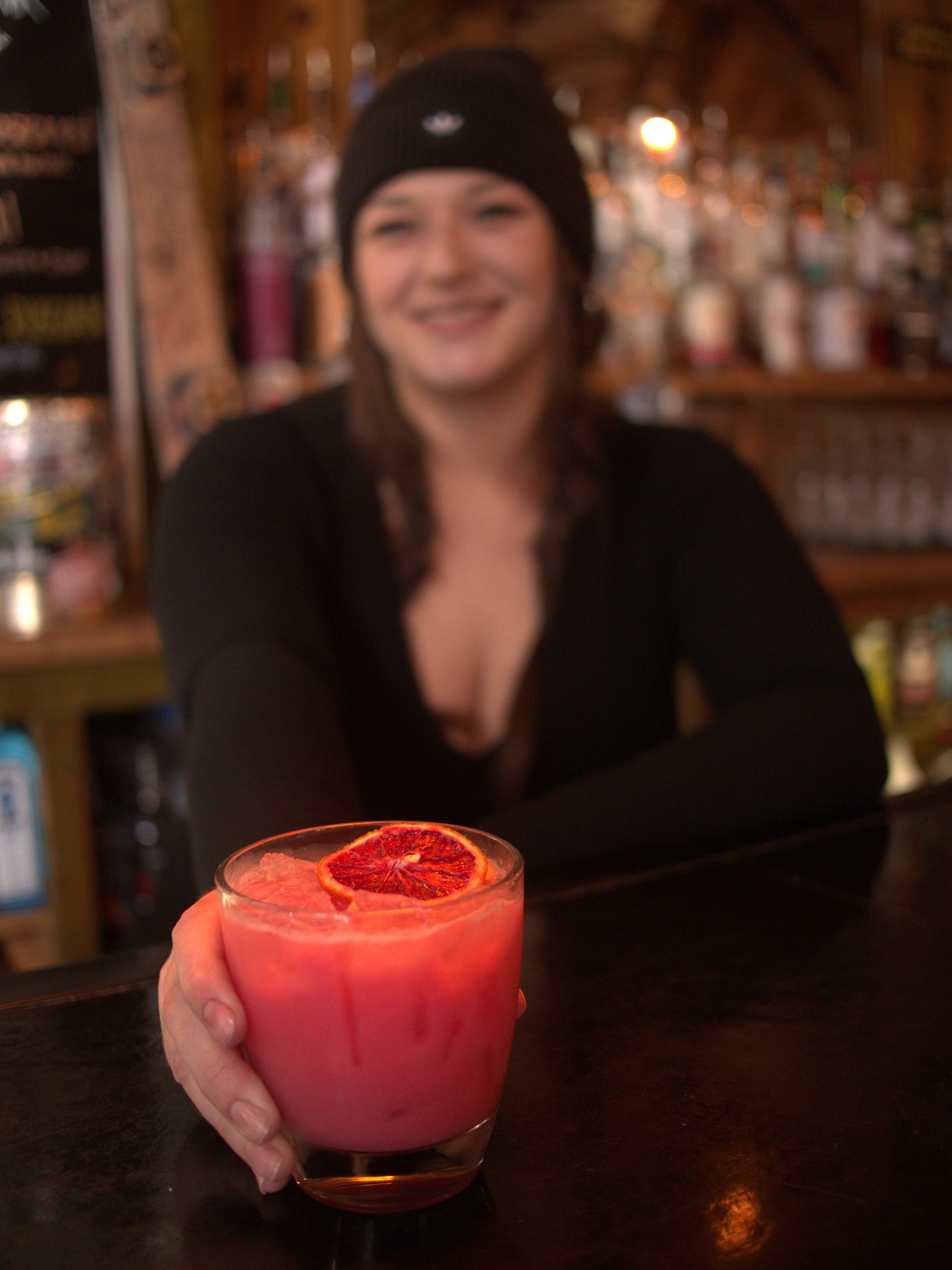 Person in a black beanie smiles at a bar, holding out a pink cocktail with a grapefruit slice.