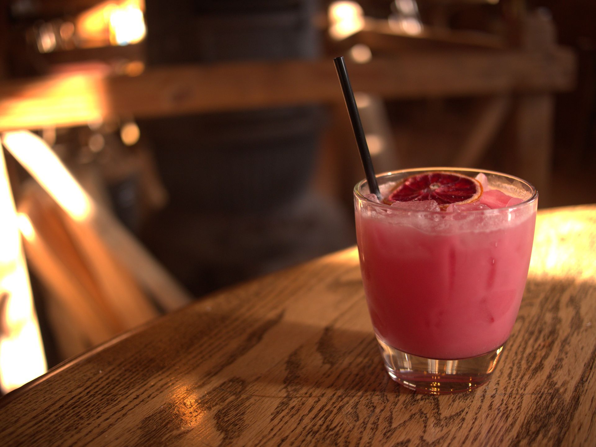 Pink cocktail with a black straw and berry garnish on a wooden table in warm bar lighting