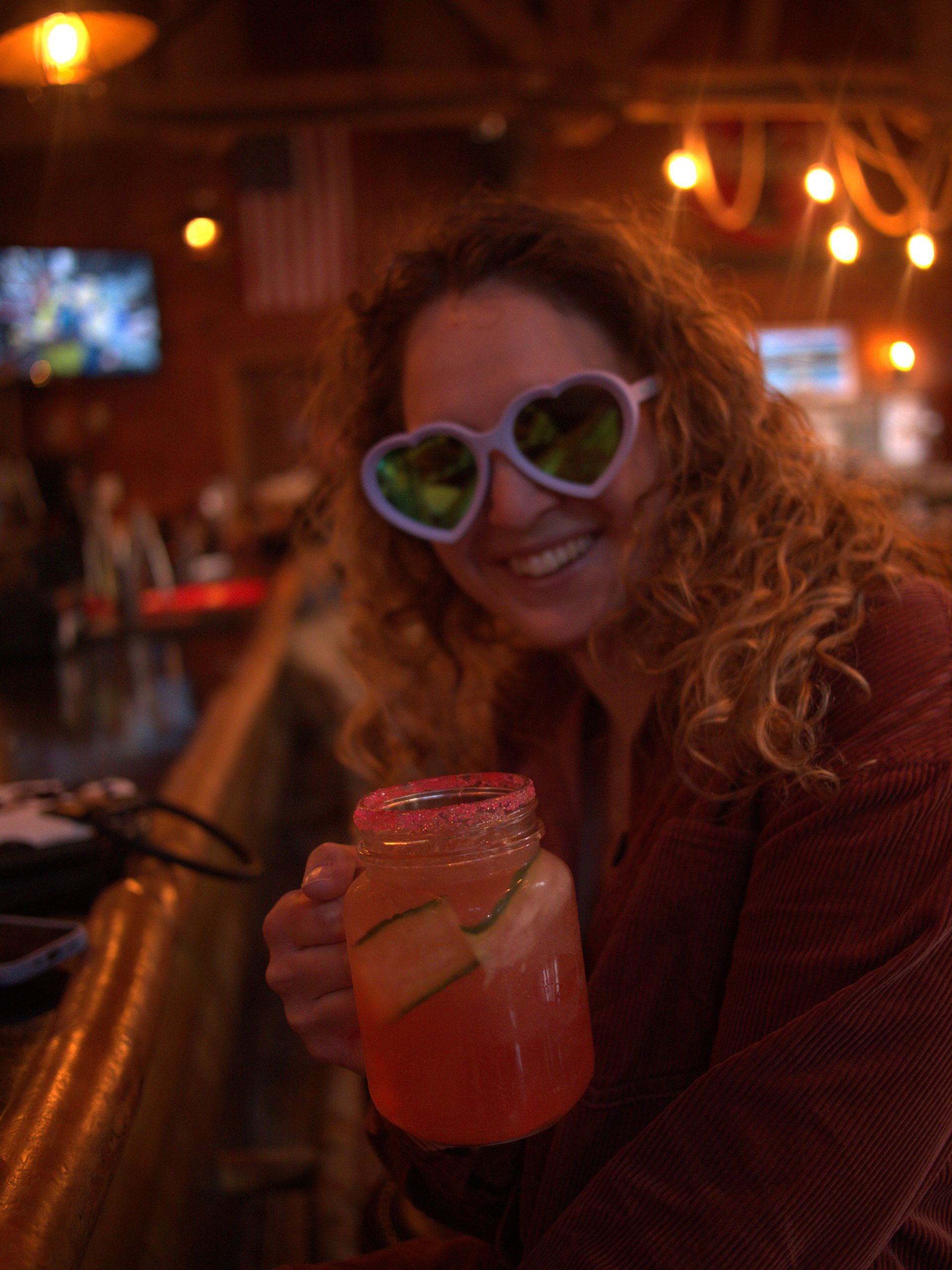 Person in heart-shaped sunglasses smiling and holding a pink drink in a warmly lit bar