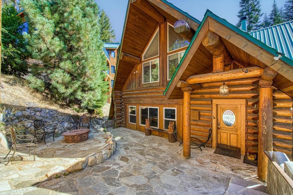 Rustic log cabin with stone patio, green roof, and pine trees in a sunny mountain setting