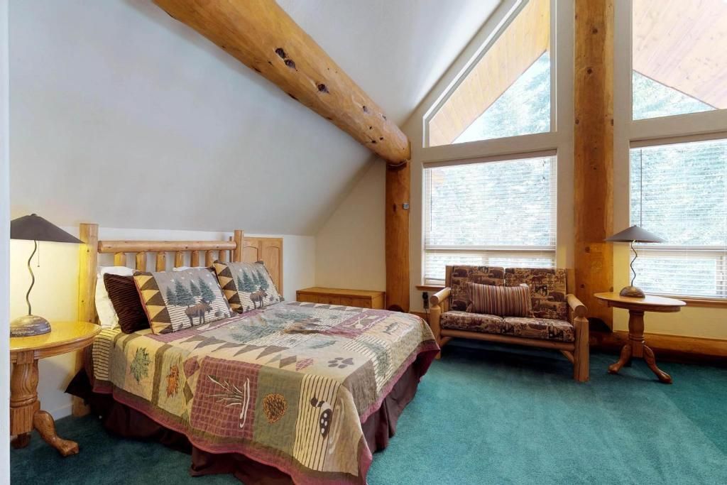Cozy attic bedroom with a bed, wicker chair, and large windows, featuring wood beams and teal carpet.