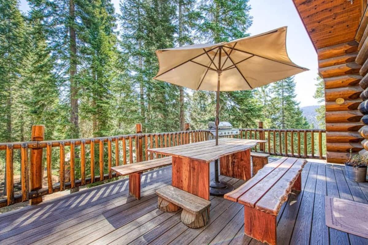 Wooden deck with picnic table, benches, and umbrella overlooking a forest of tall pine trees