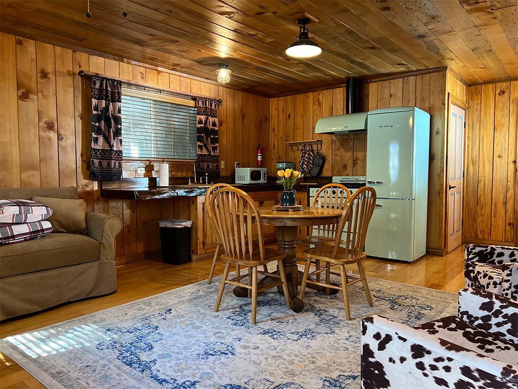 Cozy cabin interior: wood paneling, dining table, light blue rug, cow-print chair, mint fridge, and kitchen.