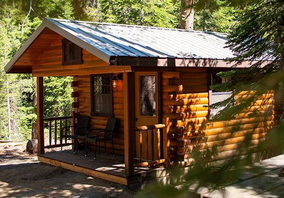 Log cabin with a porch, surrounded by trees. Exterior is wooden, roof is silver.